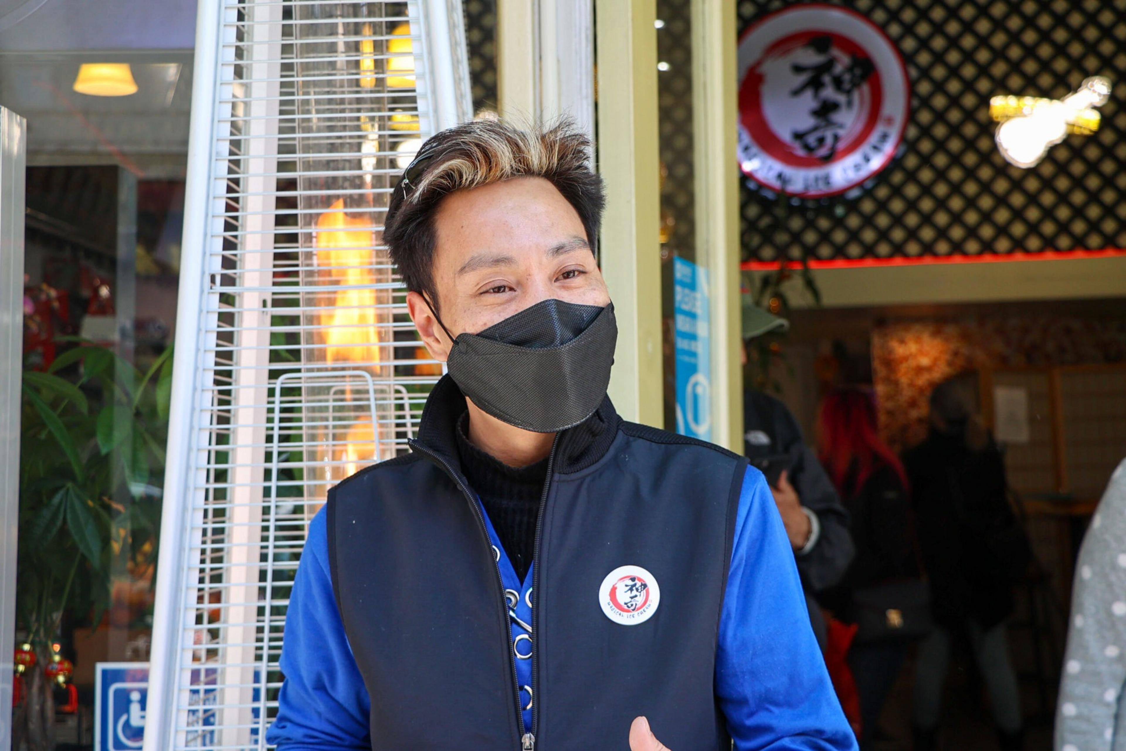 A person wearing a black face mask, blue shirt, and black vest with a round pin stands near a tall outdoor heater in front of a building with a round logo.