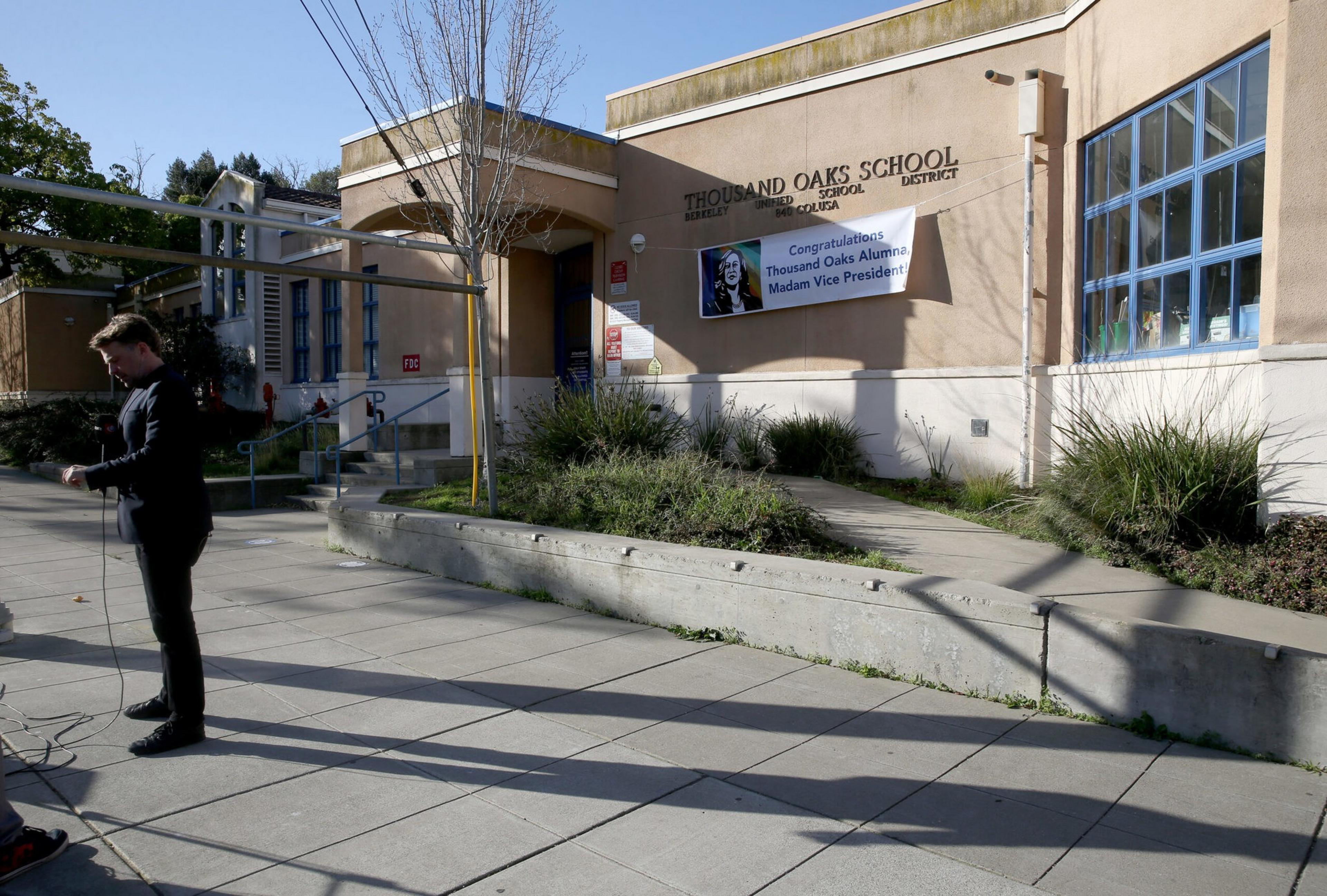 A man stands on a paved area outside Thousand Oaks School, adjusting cables, with a congratulatory banner for a Thousand Oaks alumna as Vice President on the building wall.