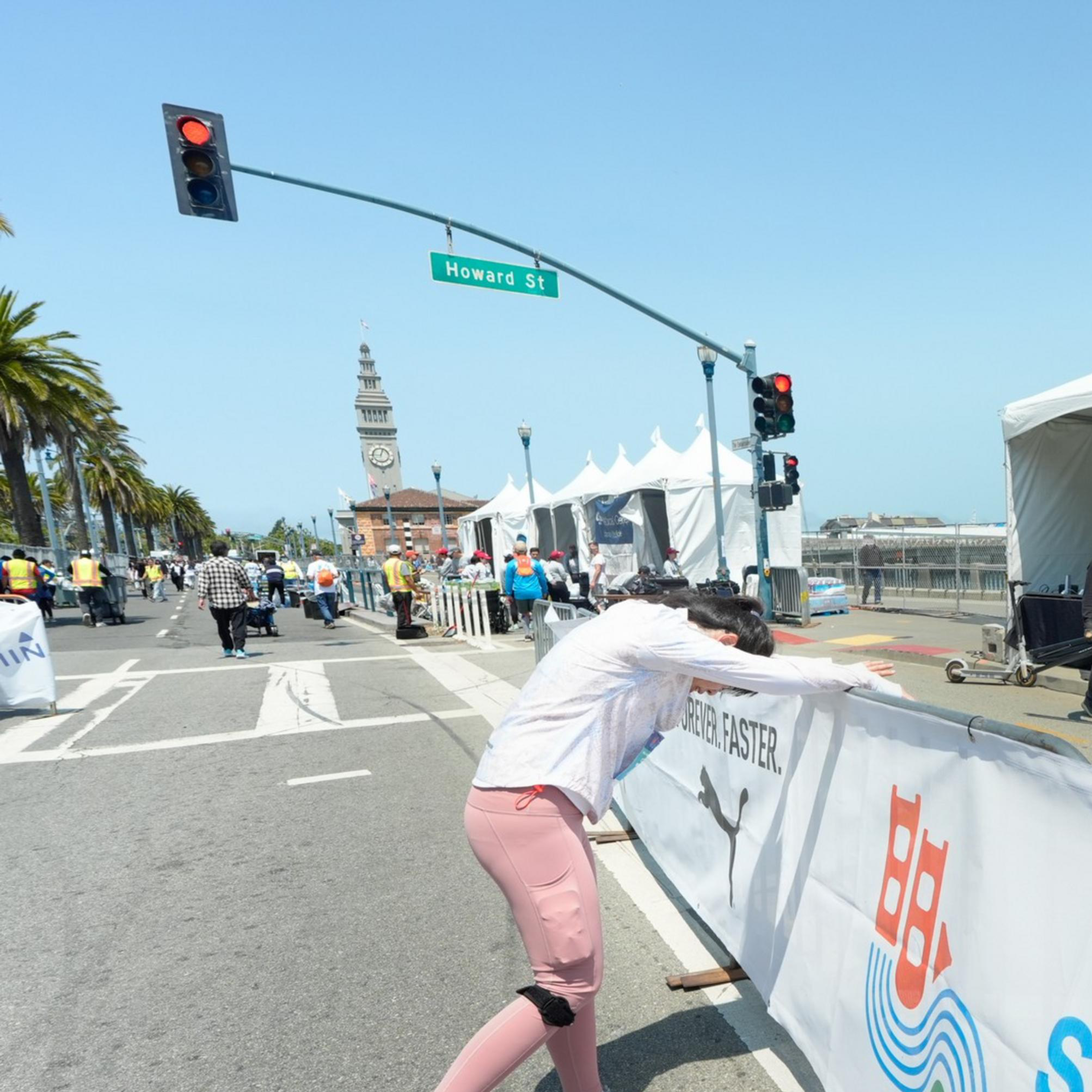 A person in pink pants leans on a barrier while a worker on a forklift carries tables. The scene is near a &quot;San Francisco Marathon&quot; sign on Howard Street.