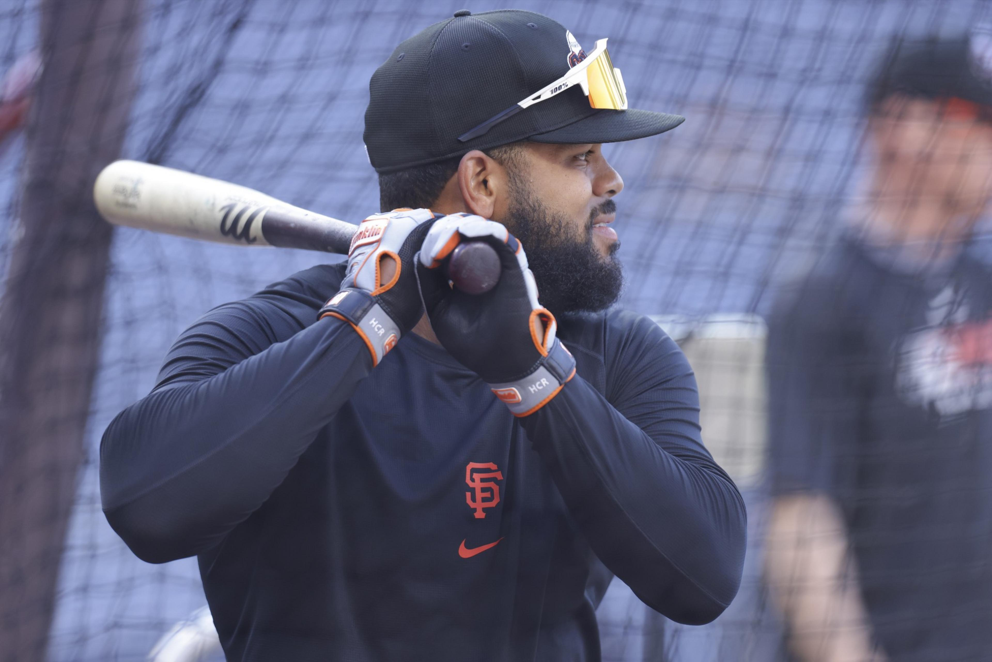 A baseball player in a black San Francisco Giants shirt and cap holds a bat, wearing batting gloves and sunglasses on his hat.