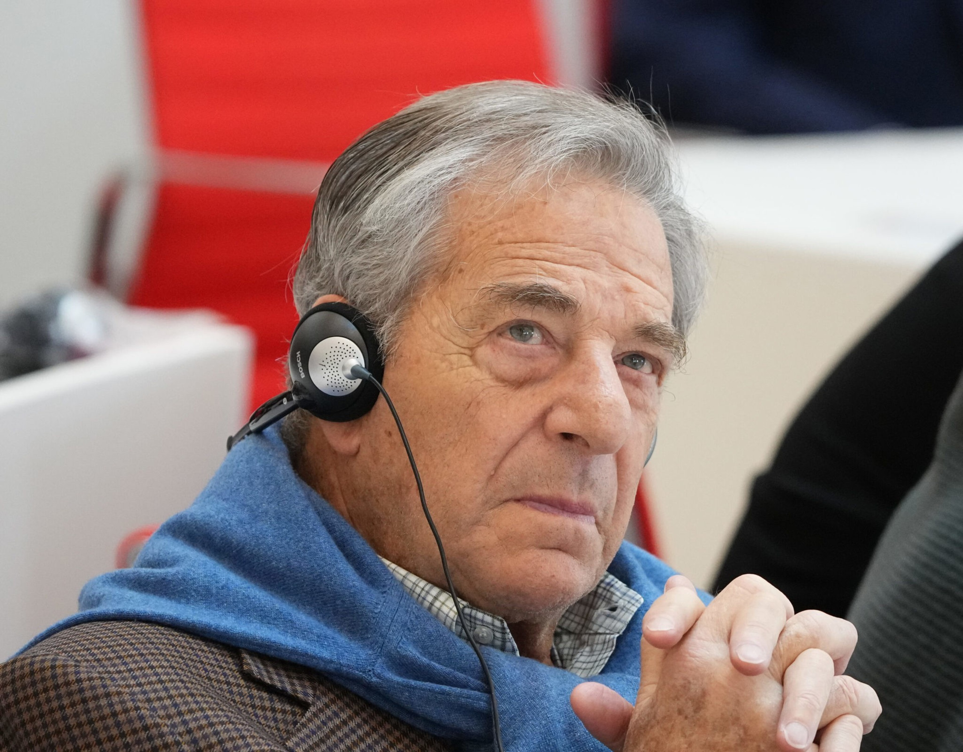 An older man with gray hair wears a blue scarf and headphones, looking thoughtfully upward with his hands clasped together.