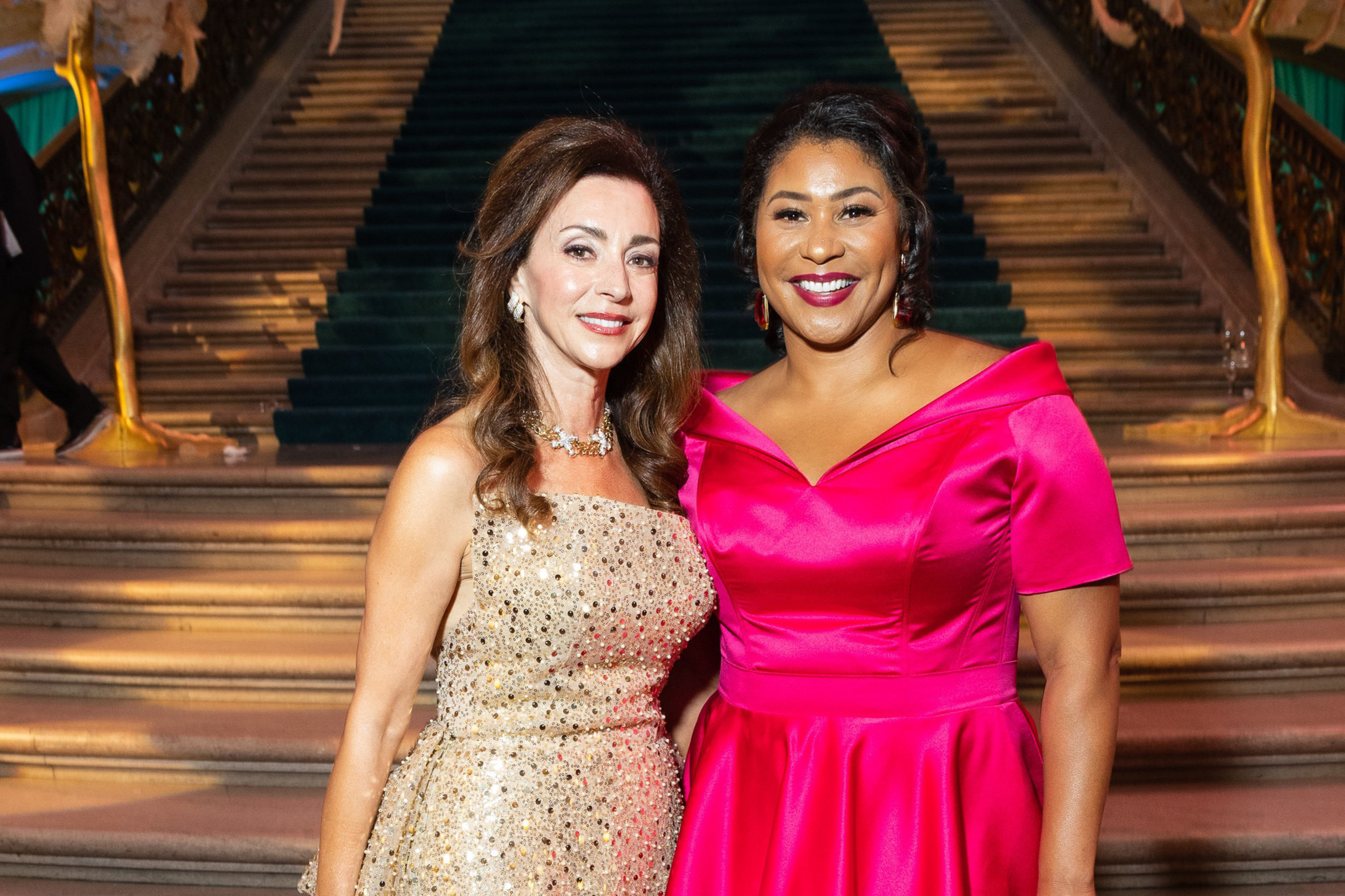 Two women stand side by side, smiling; one wears a sparkly gold dress and the other a bright pink satin dress, with stairs in the background.