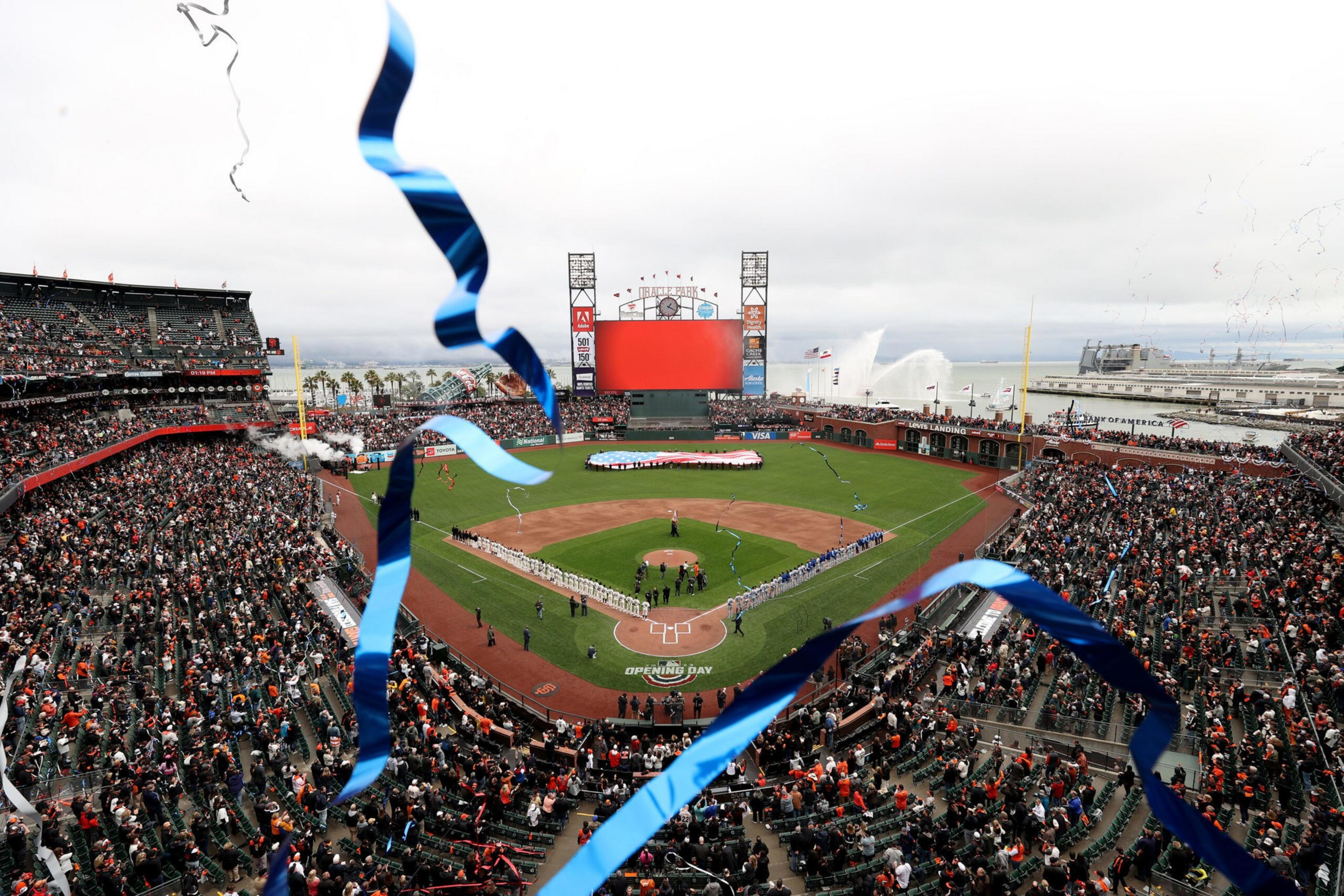 A packed baseball stadium shows teams lined up on the field with a large flag at home plate and blue streamers falling over the crowd.