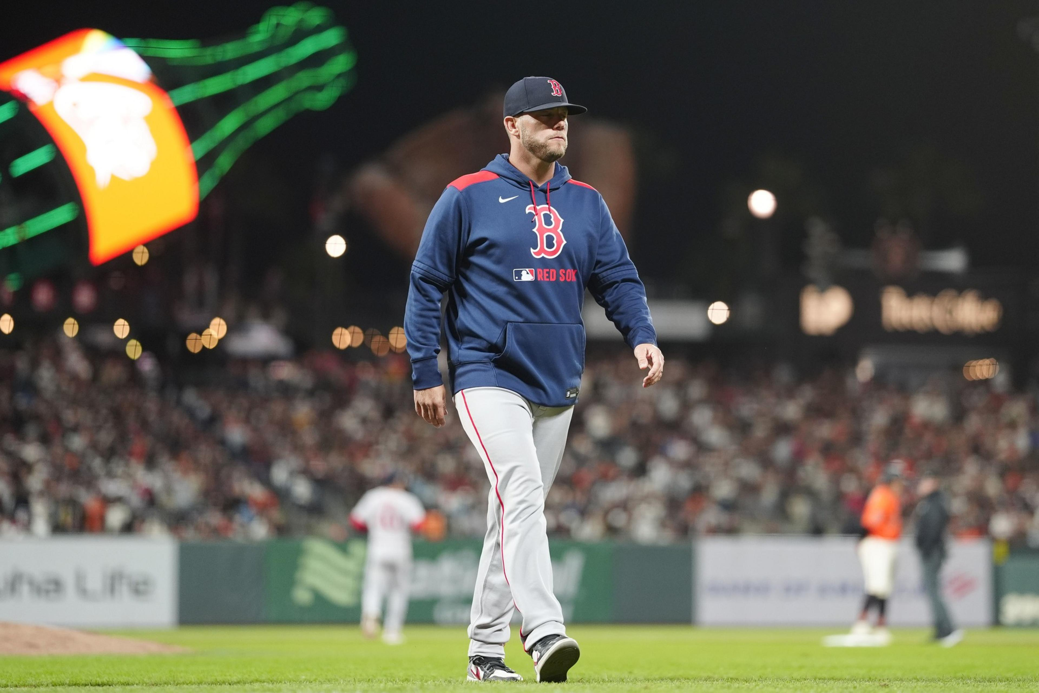 A baseball player in a Boston Red Sox jacket and cap walks on the field at night with a blurred crowd and a neon sign in the background.