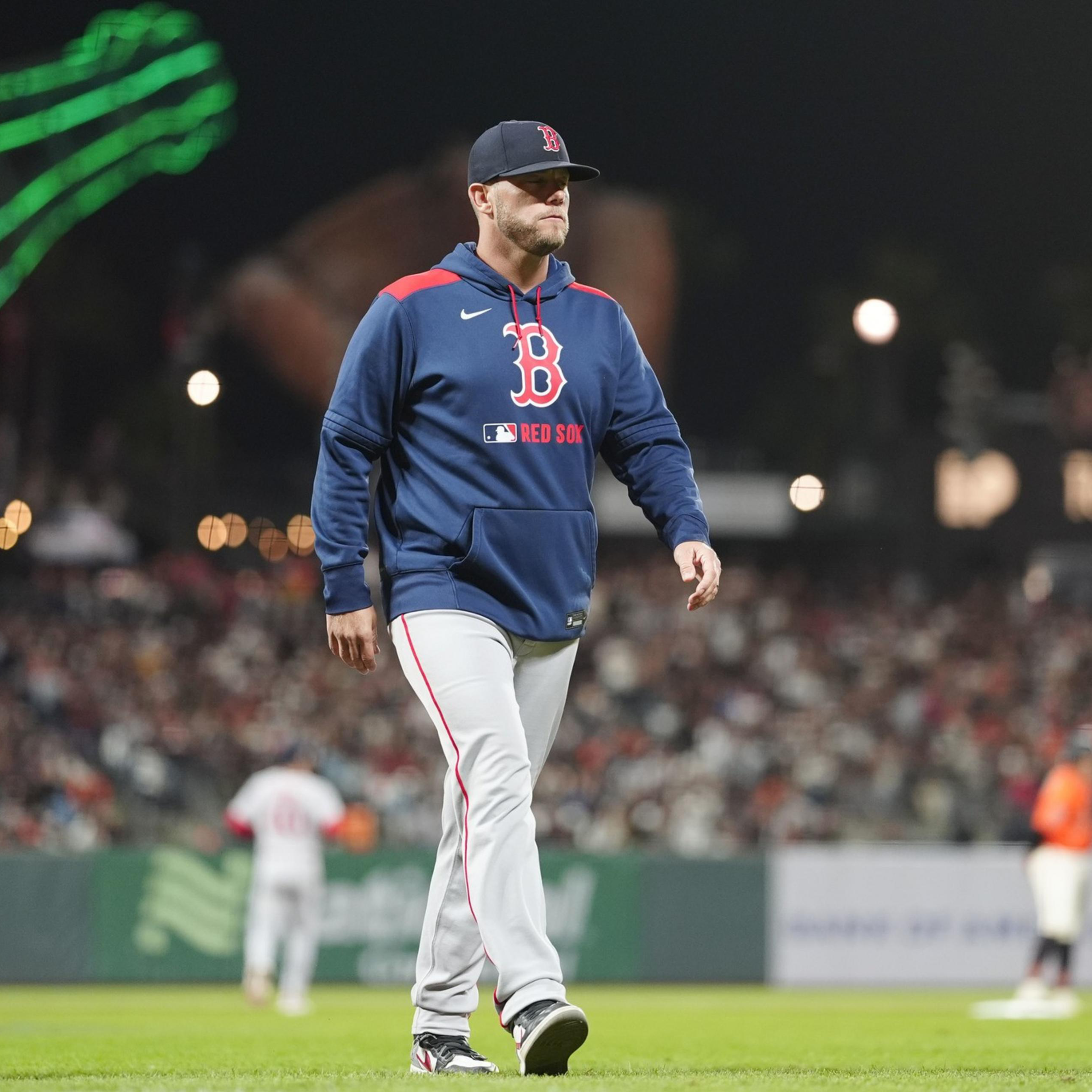 A baseball player in a Boston Red Sox jacket and cap walks on the field at night with a blurred crowd and a neon sign in the background.
