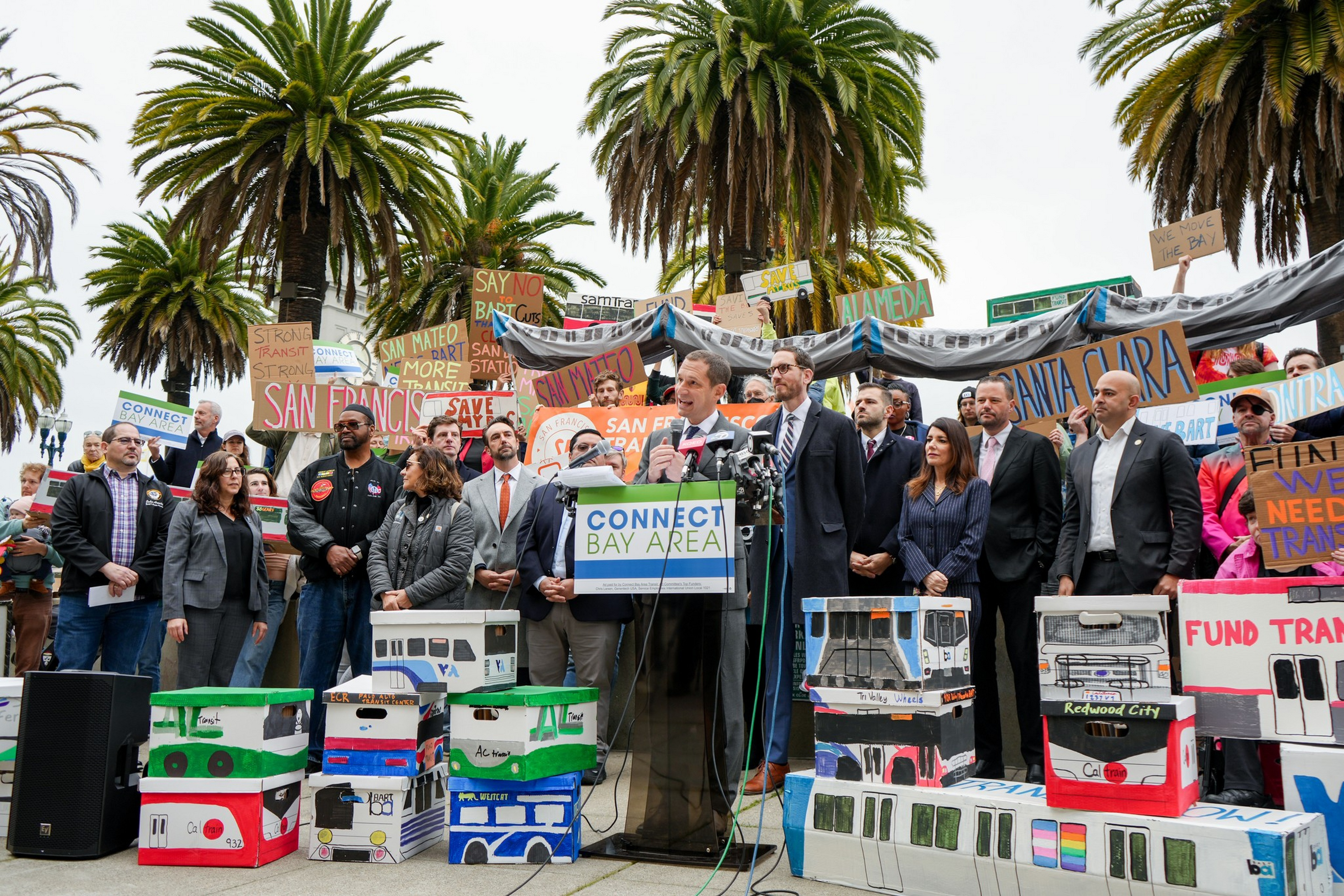 A group of people stand by palm trees holding signs advocating for Bay Area transit funding, with speakers at a podium labeled "Connect Bay Area" and model trains displayed.