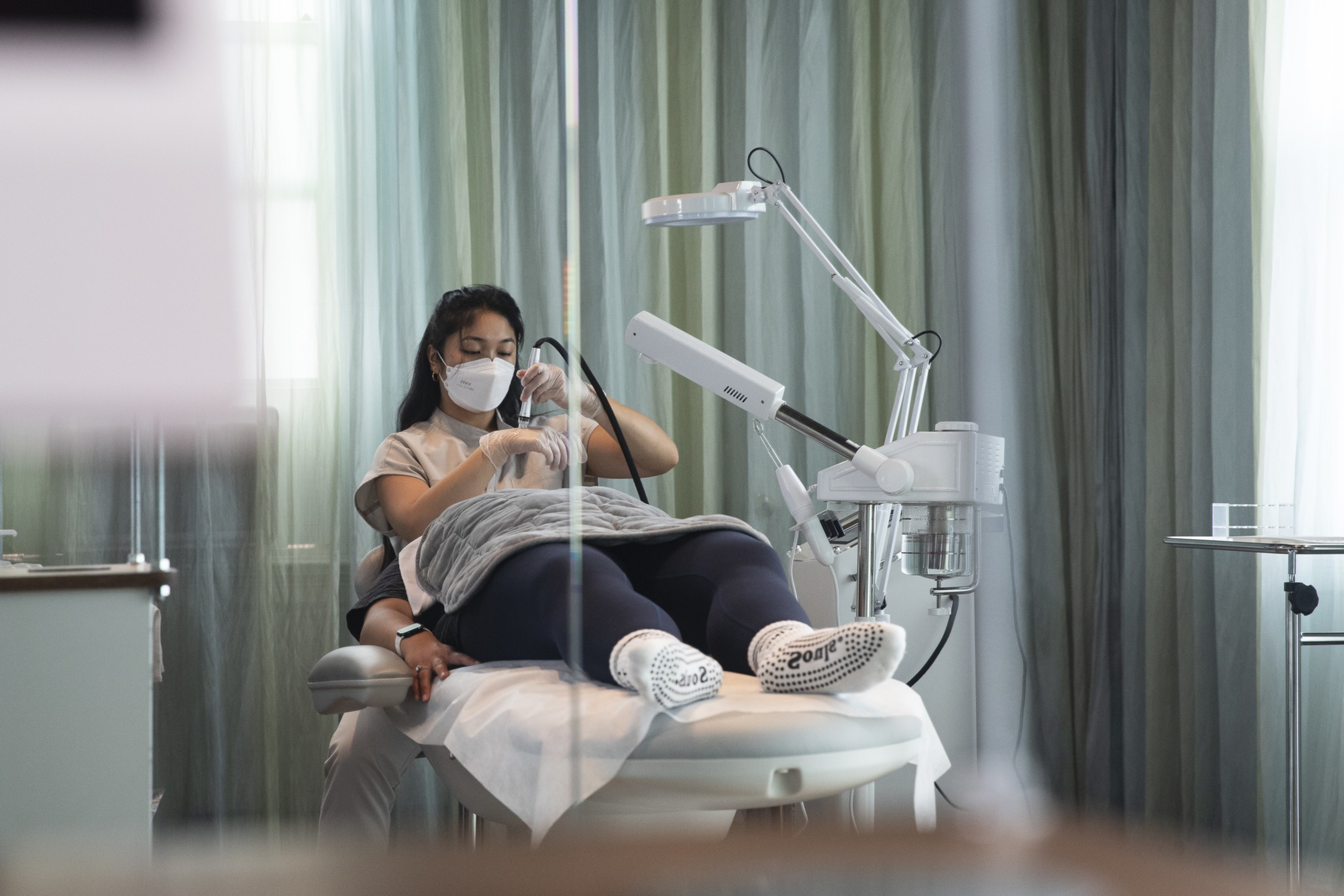 A woman wearing a mask lies on a treatment bed while a masked esthetician performs a facial procedure using a handheld device.