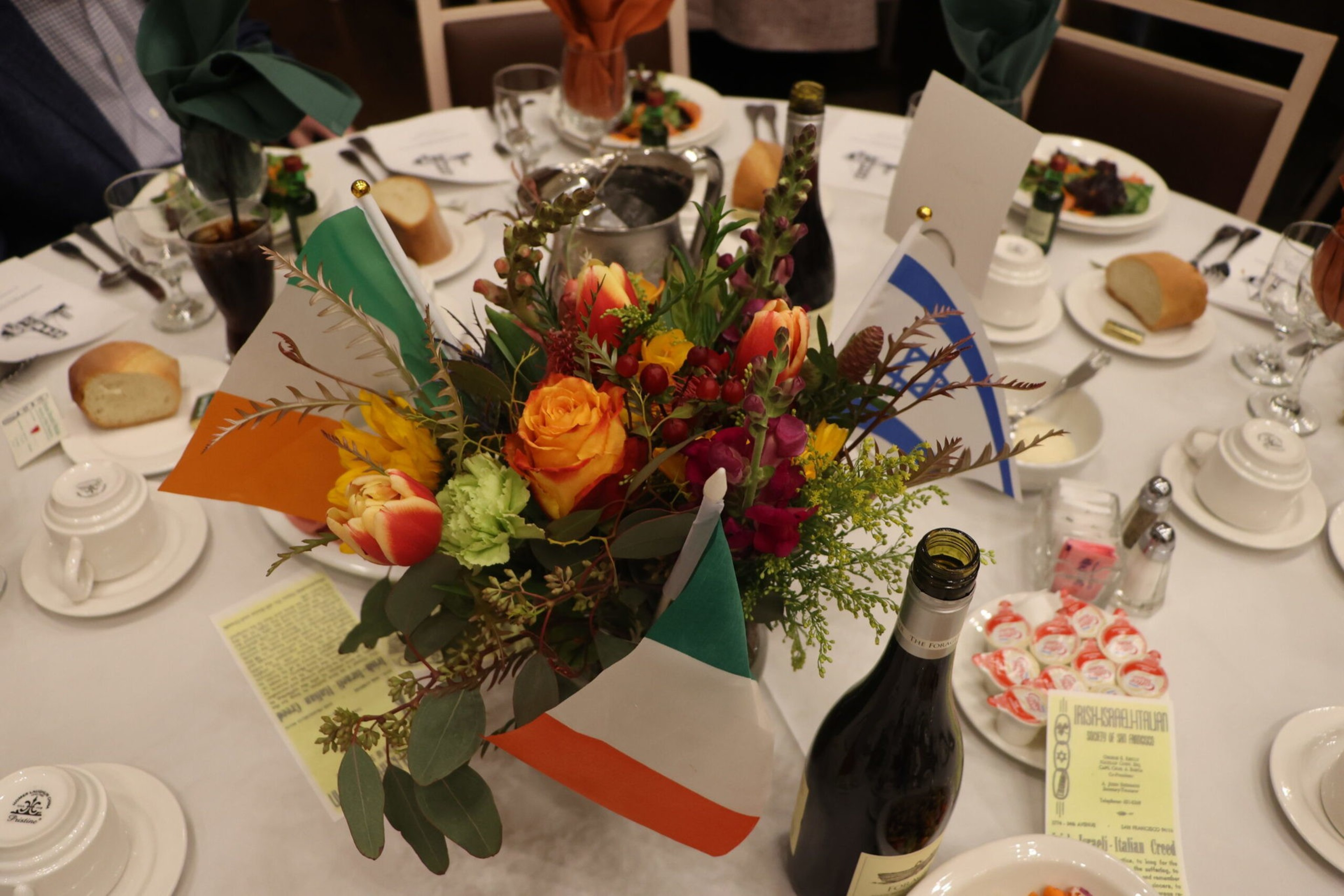 A round banquet table set with white china, bread rolls, wine, soda, floral centerpiece with orange roses, tulips, greenery, and small flags including the Irish flag.