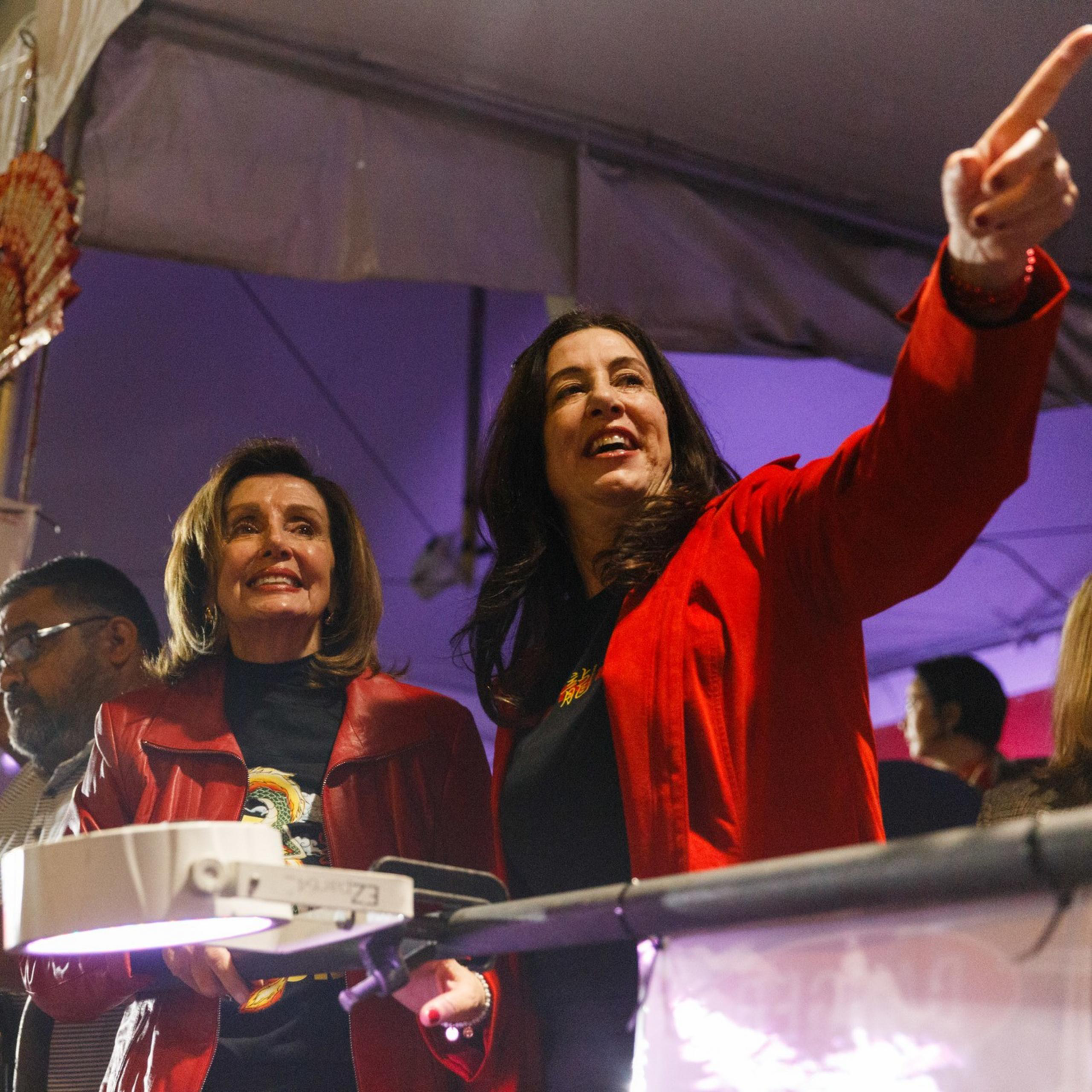 Two women in red jackets appear cheerful at an event, one pointing excitedly, surrounded by onlookers with cameras.