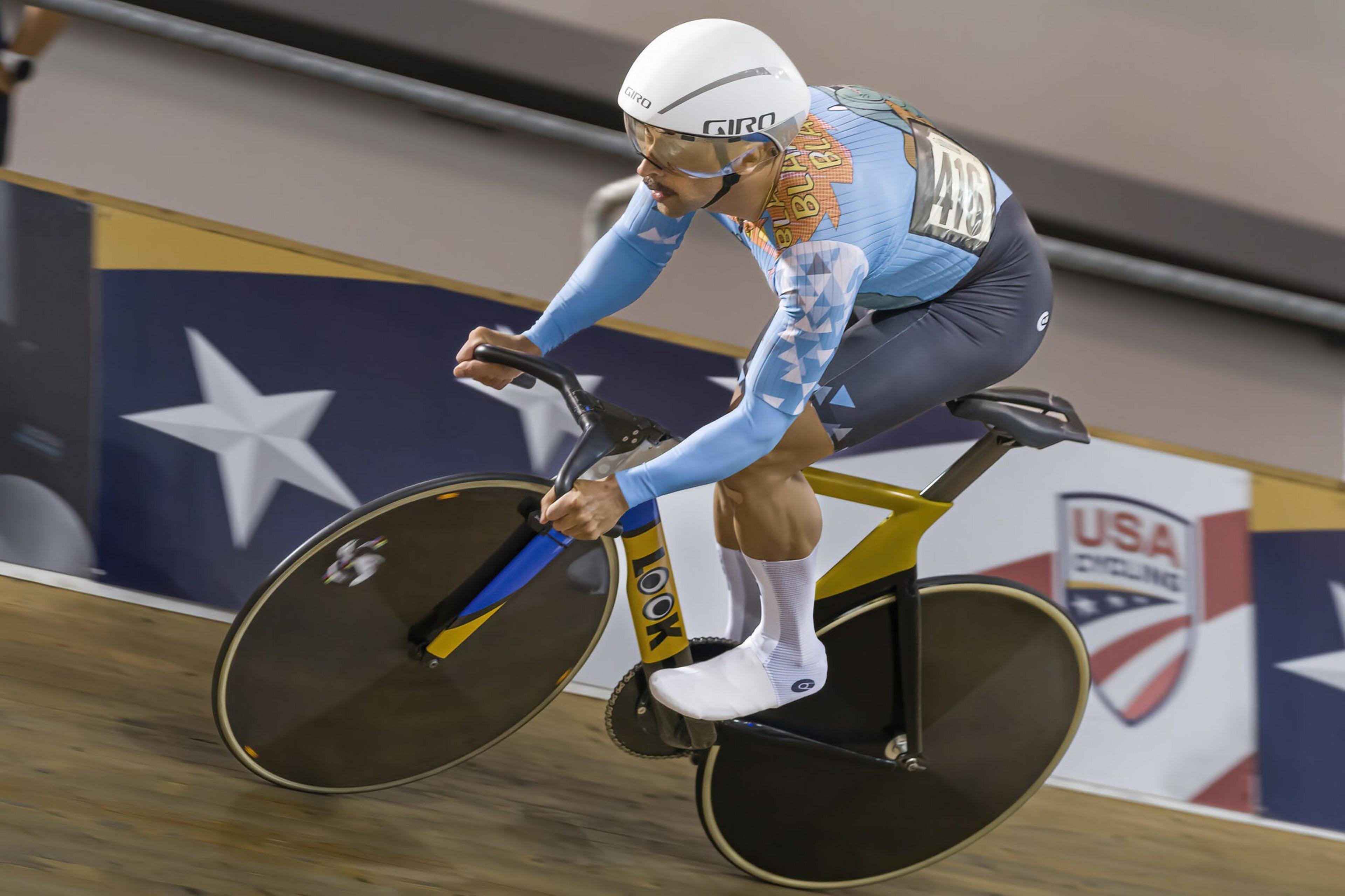 A cyclist wearing a blue and gray kit with a white helmet and socks rides a yellow and black bike on an indoor track, with USA Cycling logos visible in the background.