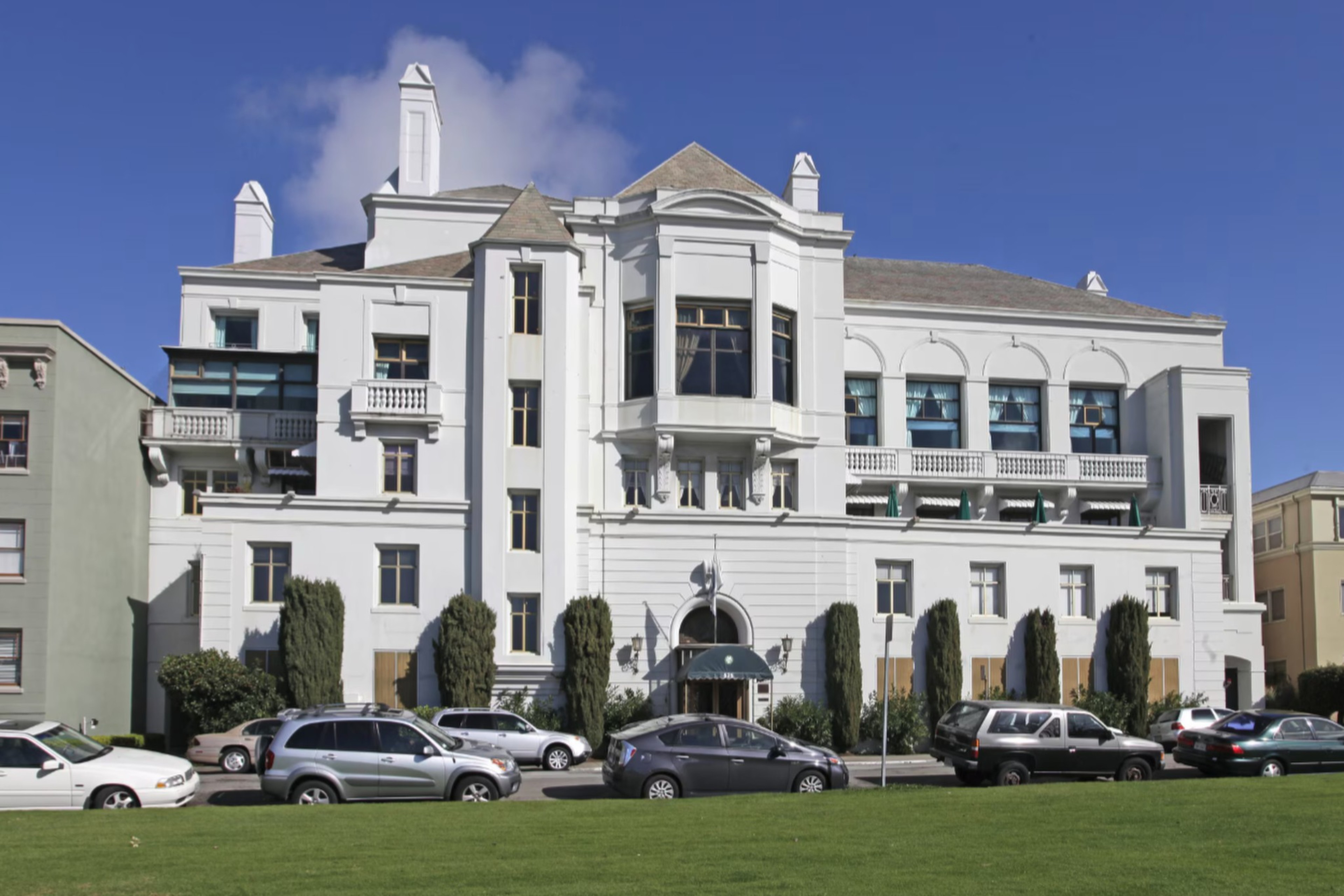 A large white multi-story building with classic architectural details stands behind a row of parked cars and manicured bushes under a clear blue sky.