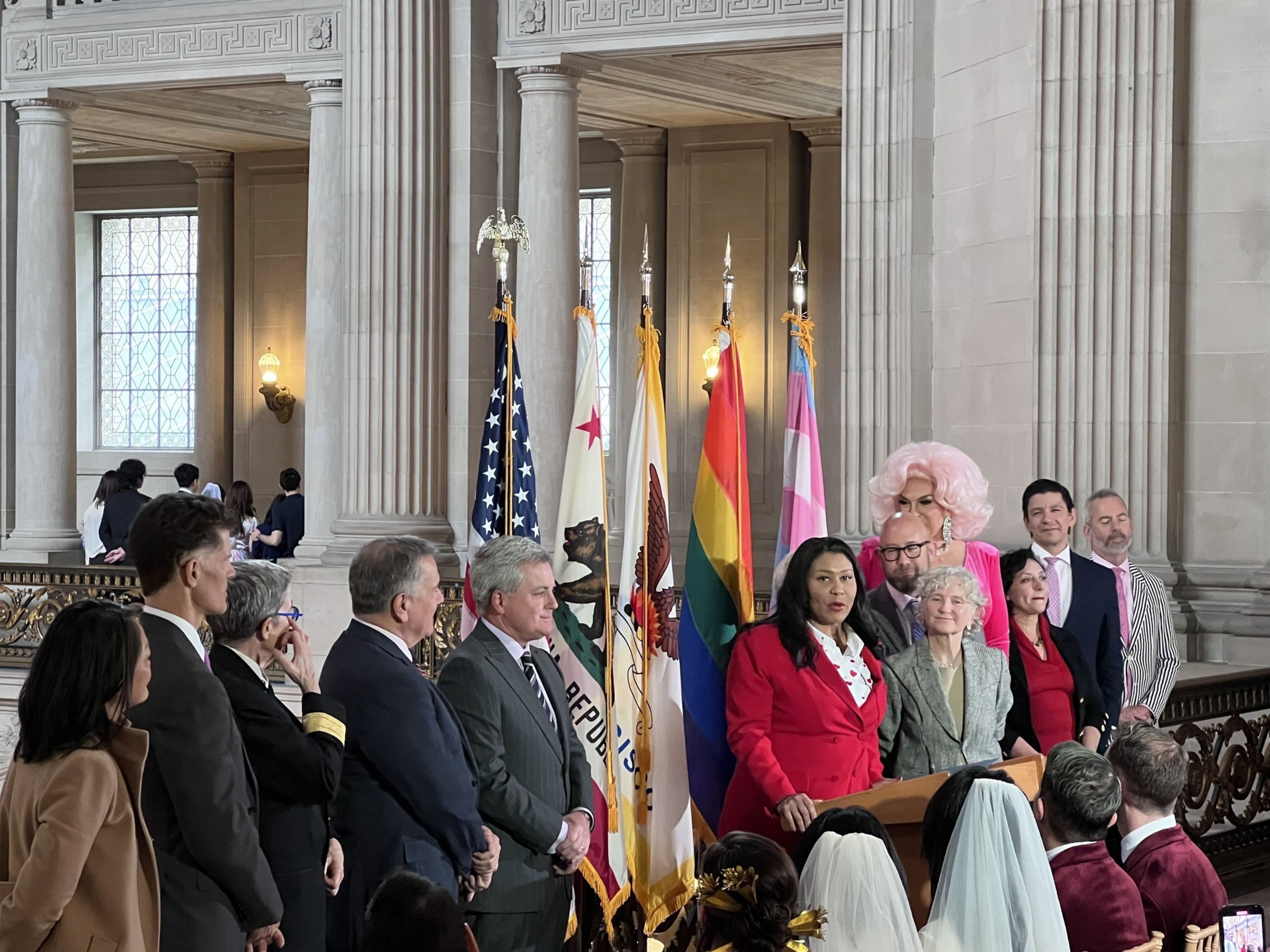 Eleven city officials stand in front of bright flags on a balcony in San Francisco City Hall. Mayor London Breed is speaking at a podium.
