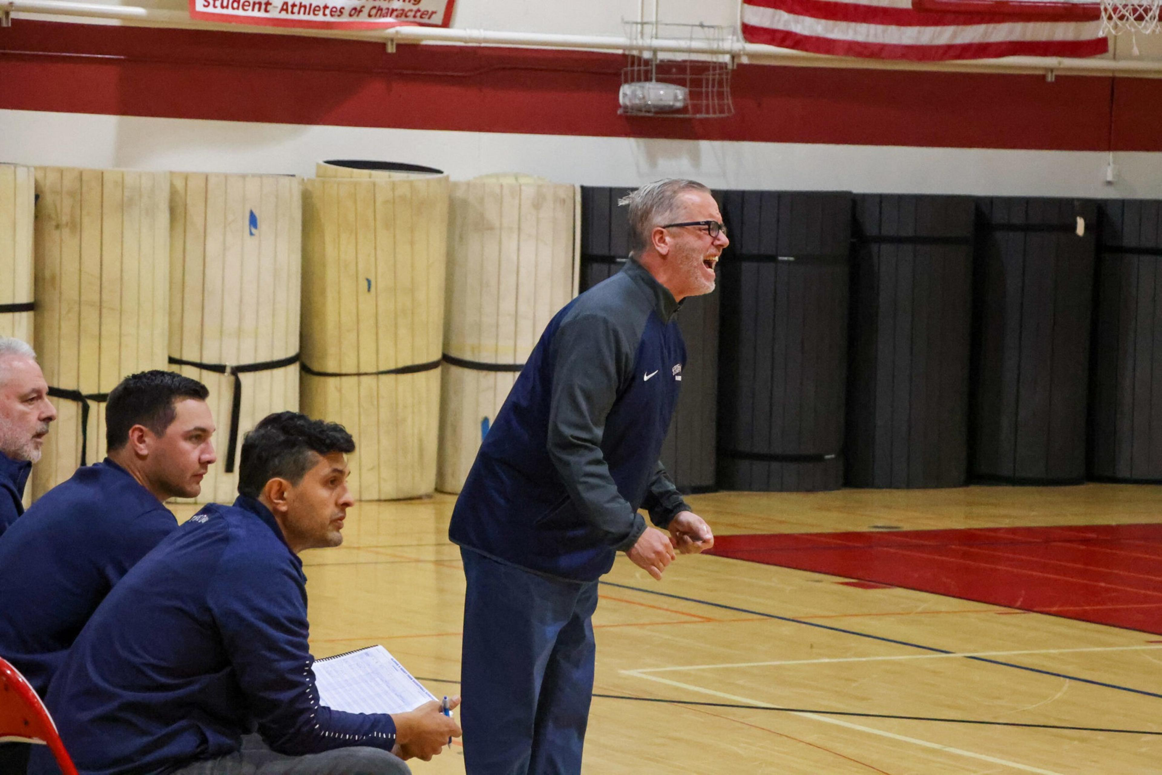 A man in glasses and blue sportswear is standing and shouting on a gym court, while three seated men nearby watch intently, one holding a clipboard.