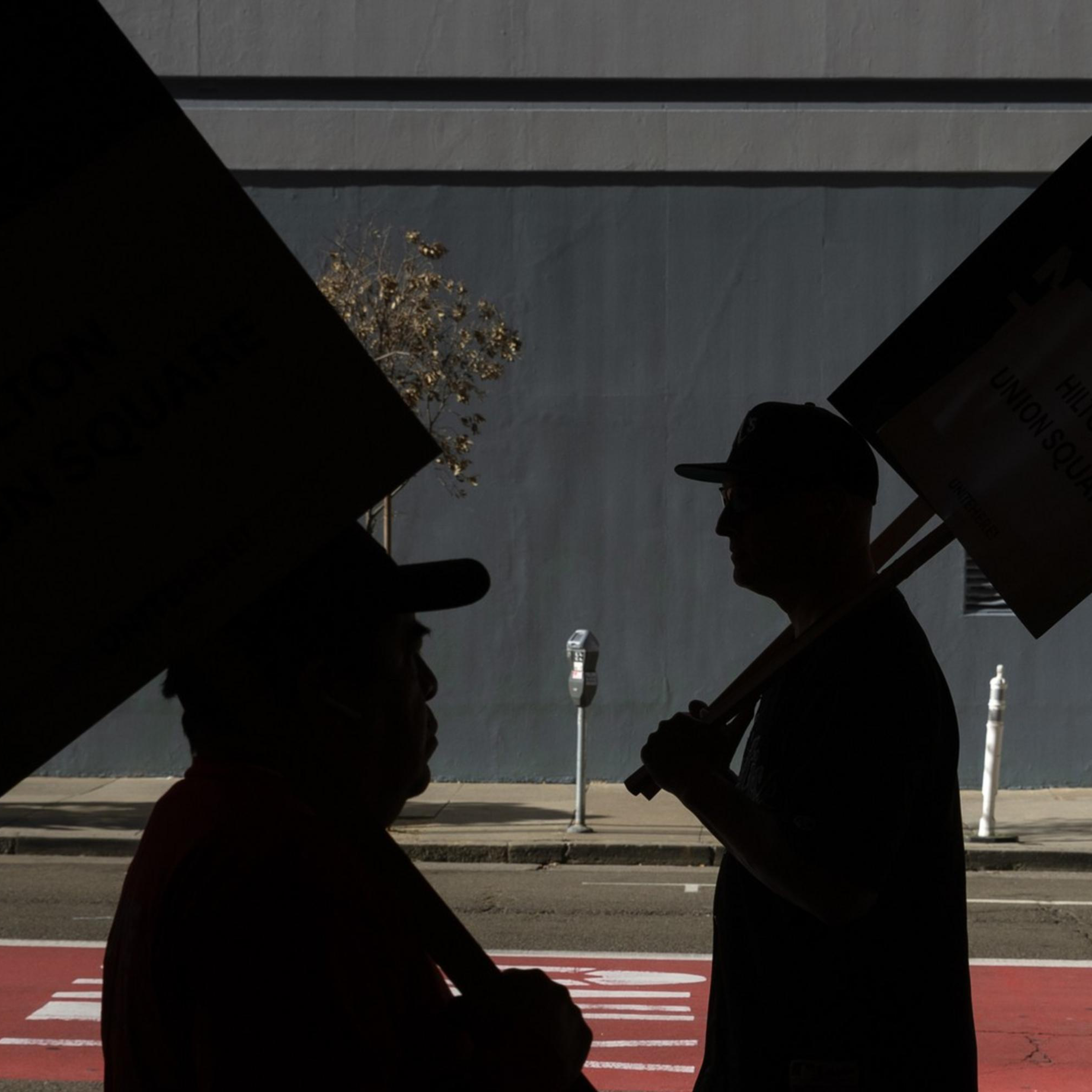 People carrying "On Strike" signs walk along a sidewalk, their silhouettes stark against the backdrop of a gray building and an empty street.