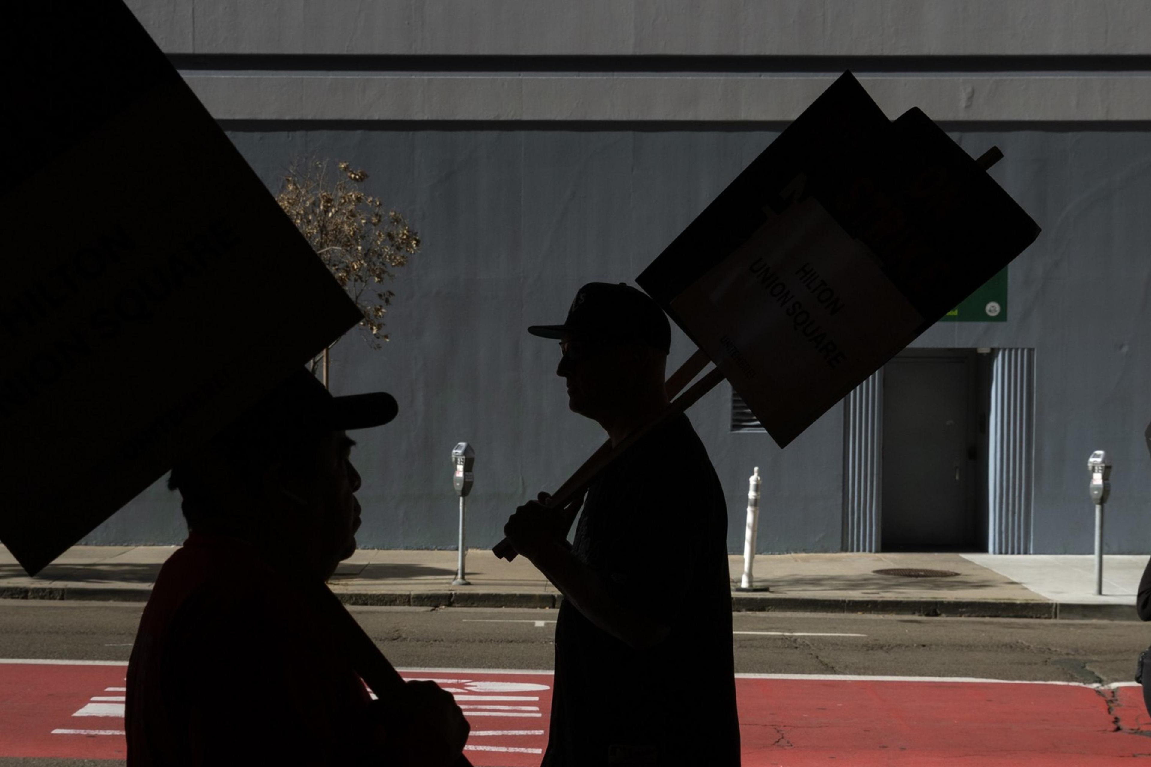 People carrying "On Strike" signs walk along a sidewalk, their silhouettes stark against the backdrop of a gray building and an empty street.