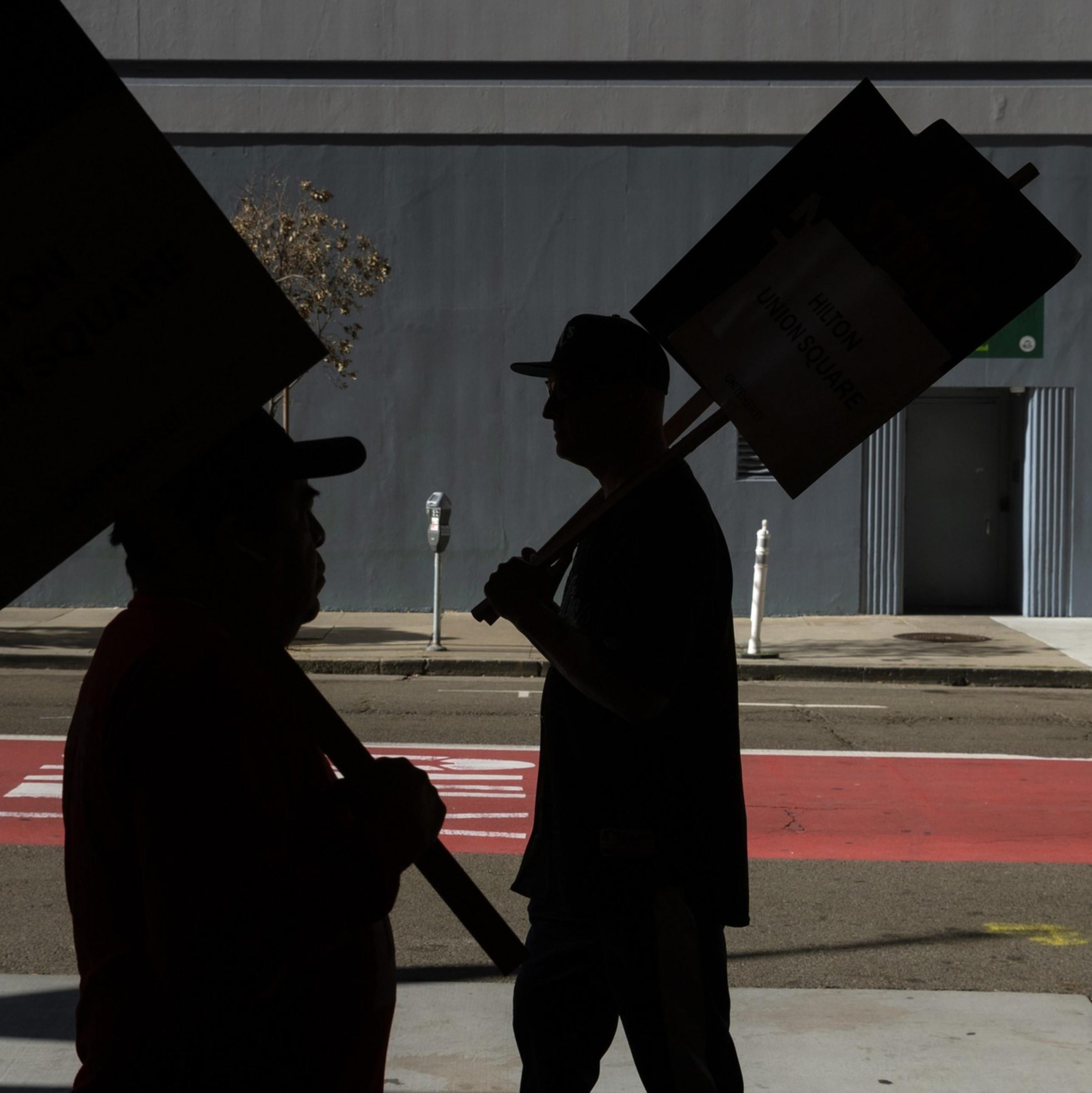 People carrying "On Strike" signs walk along a sidewalk, their silhouettes stark against the backdrop of a gray building and an empty street.