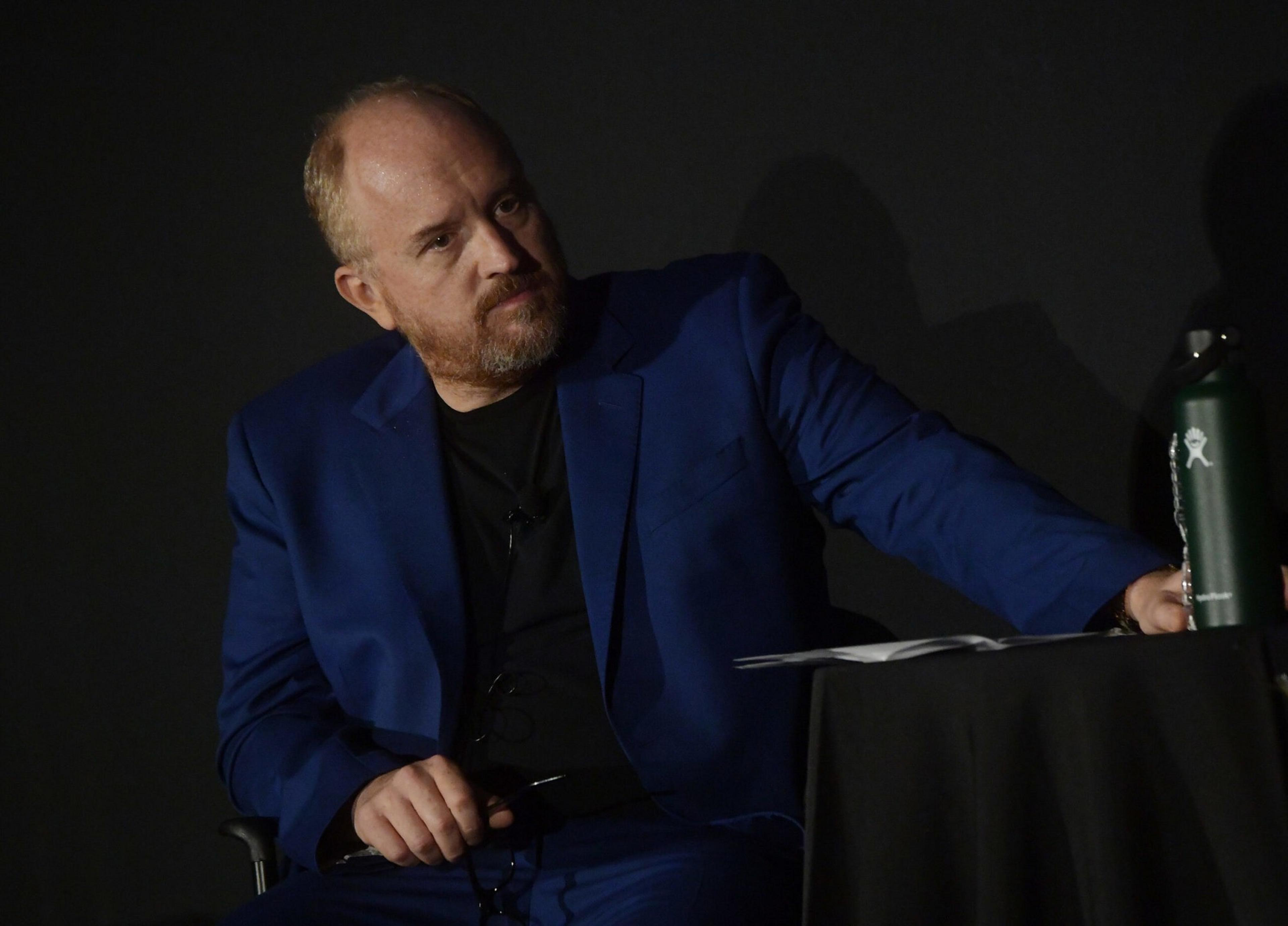 A man in a blue suit and black shirt sits at a table with papers, reaching for a green water bottle, against a dark background.