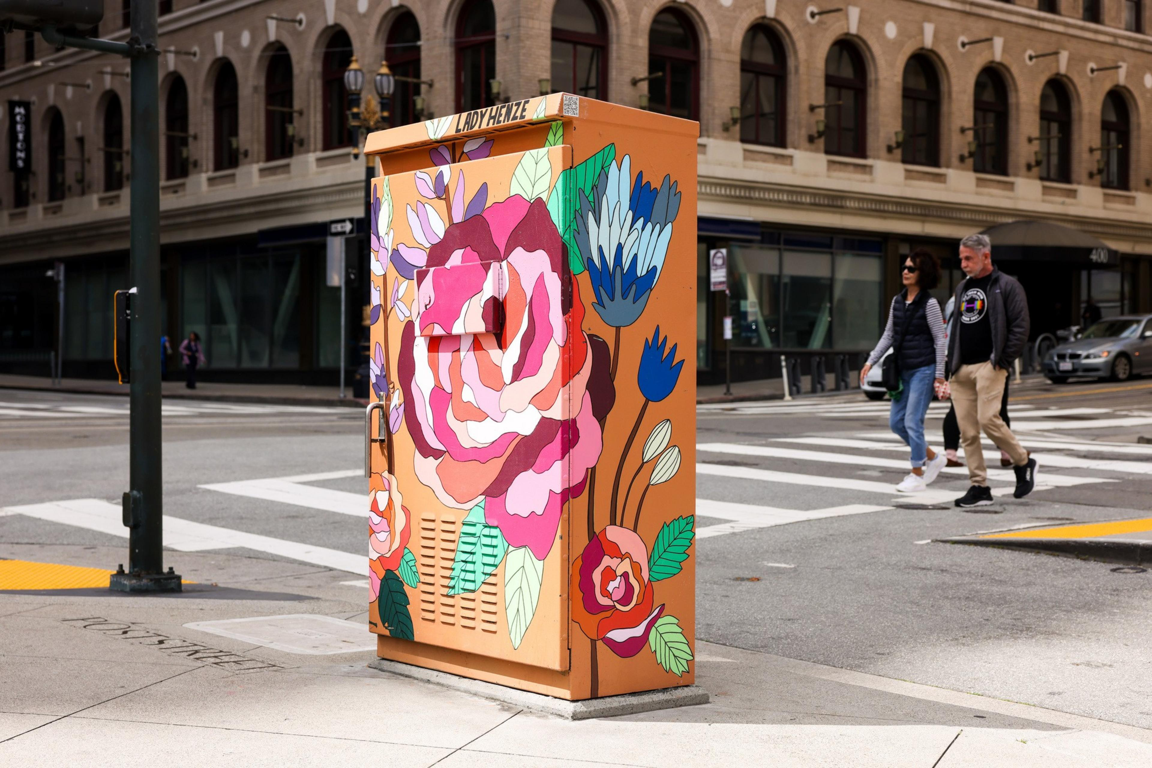 A city utility box painted with large, colorful flowers is situated on a street corner. A couple walks by, and a building with arched windows is in the background.
