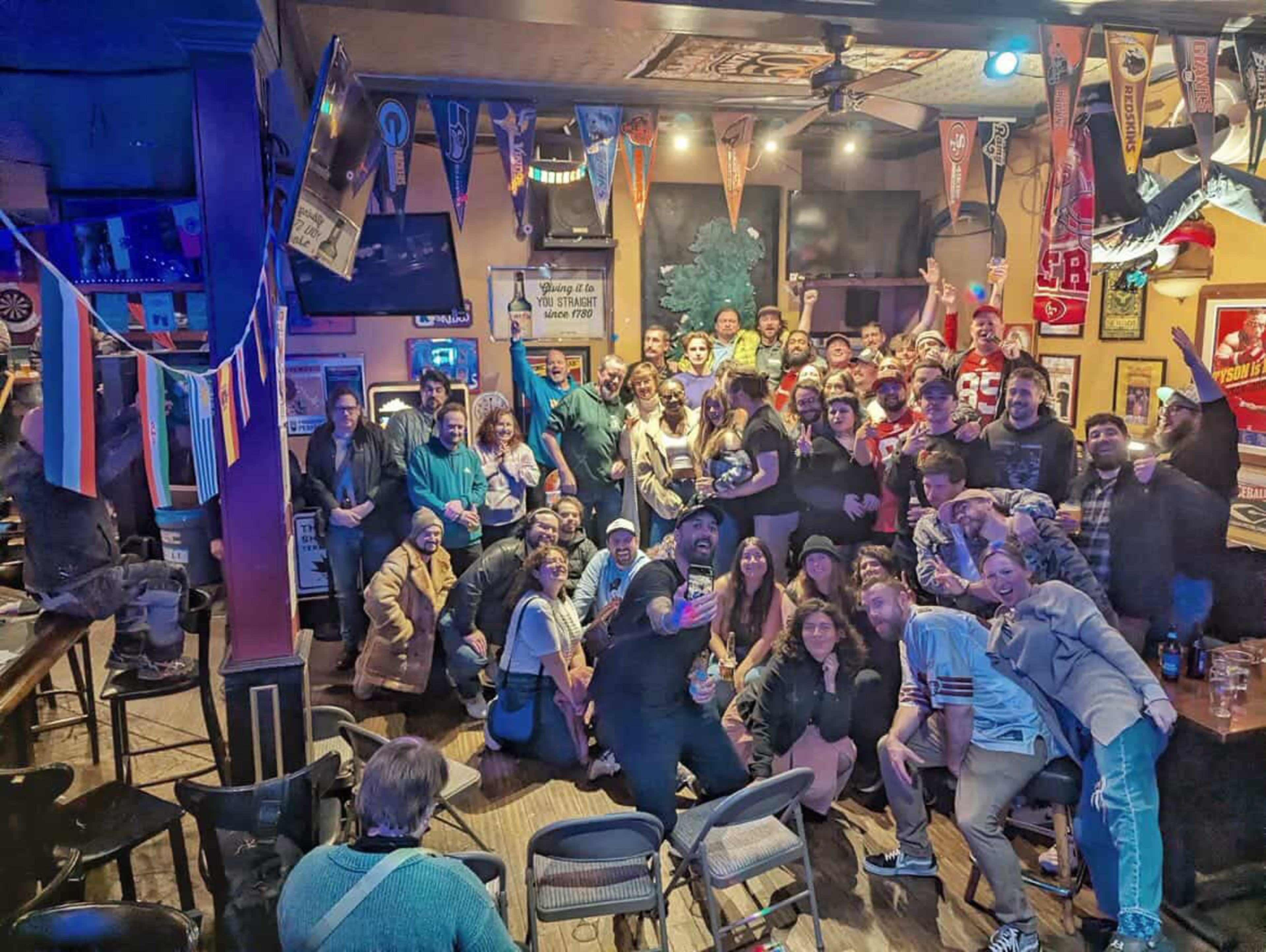 A large group of people pose closely together in a lively, decorated bar with sports banners and dim lighting.