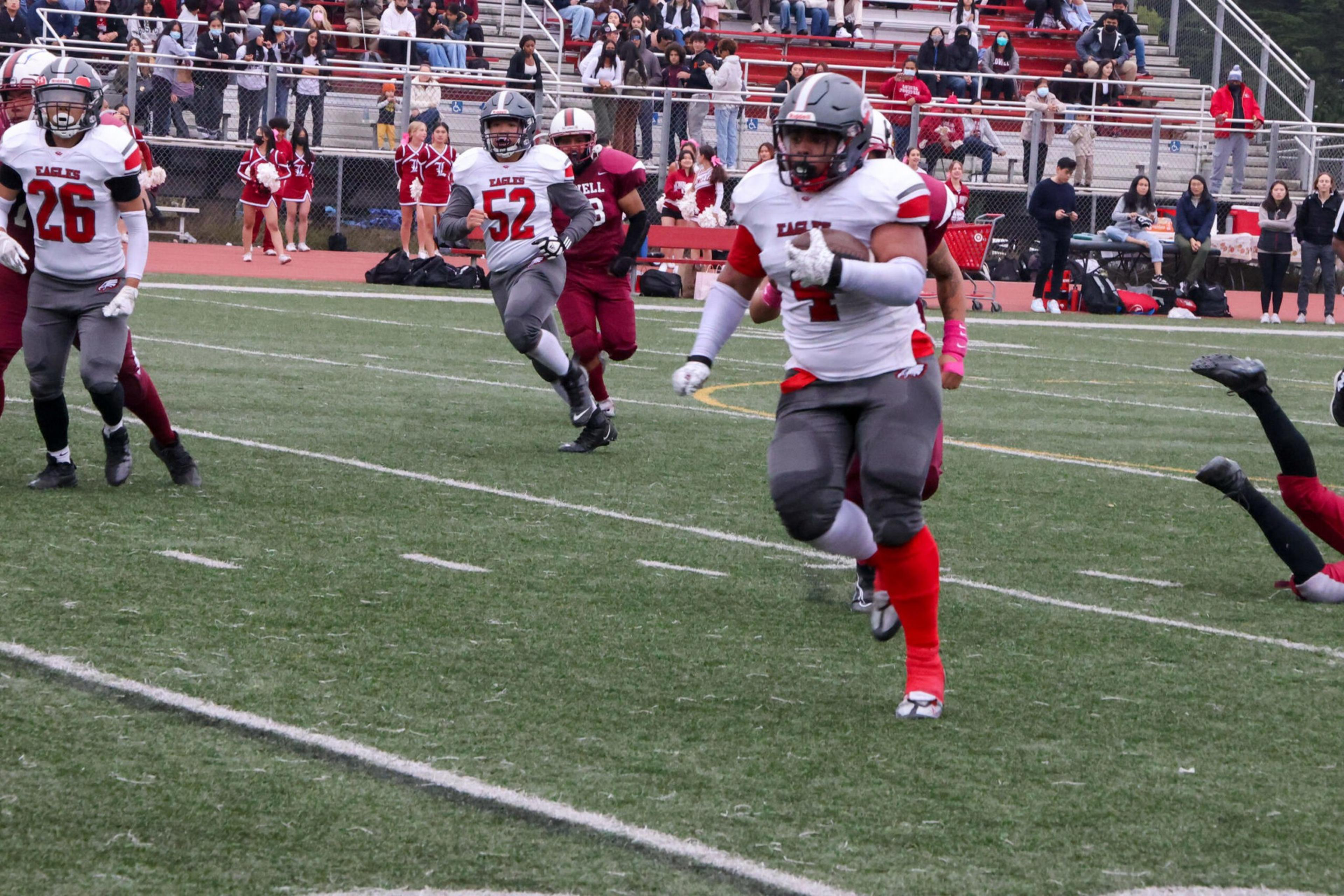 A football player in a white and gray uniform runs with the ball while opponents and teammates chase him on a green field with spectators in the background.