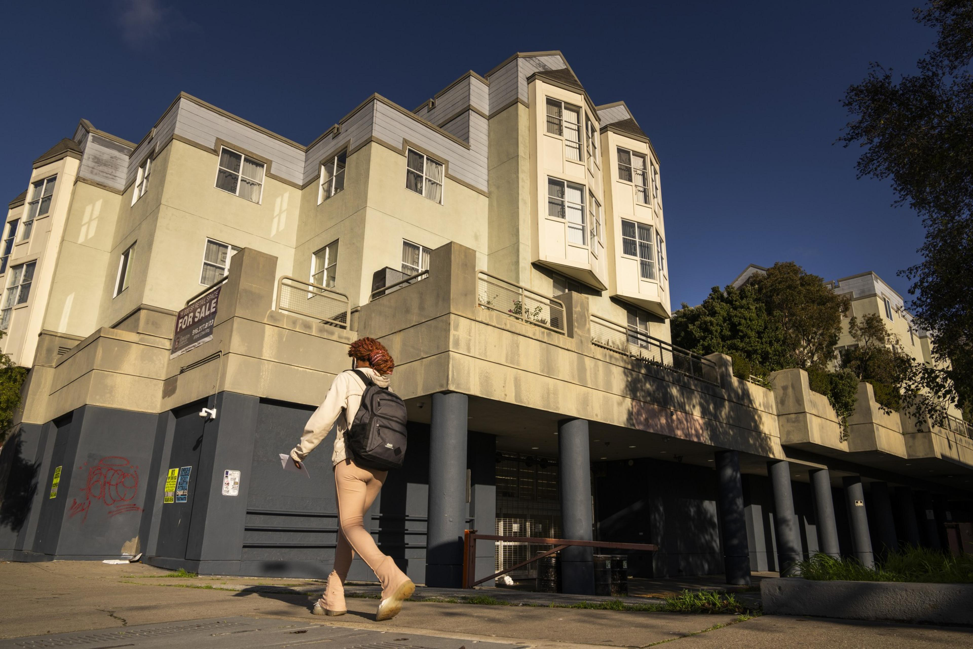 A person walks by a beige urban building with a ""For Sale"" sign under a sunny sky.