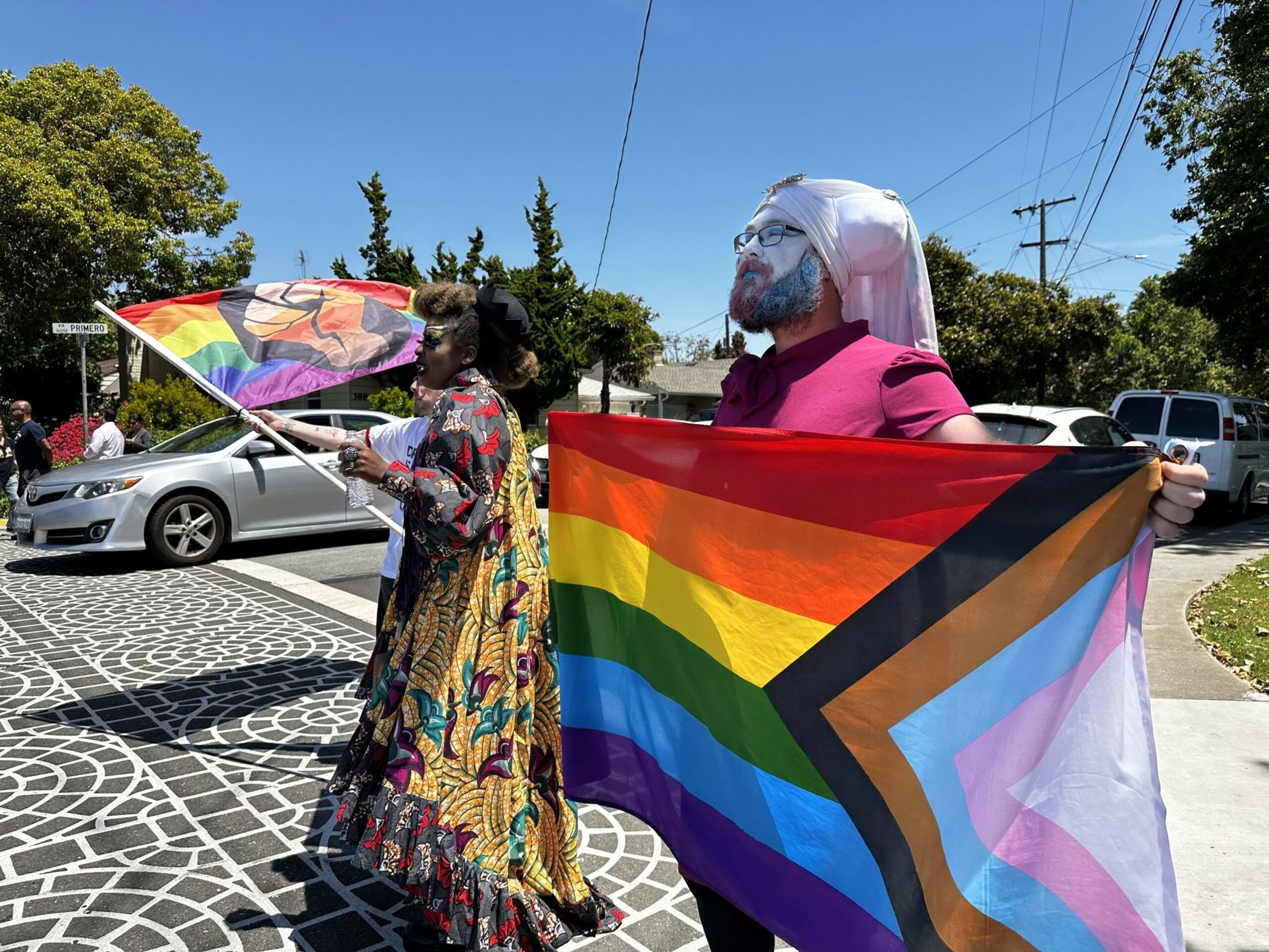 Two people stand outdoors on a sunny day holding pride flags, one with a raised fist symbol and the other a Progress Pride flag with black, brown, blue, pink, and white stripes.