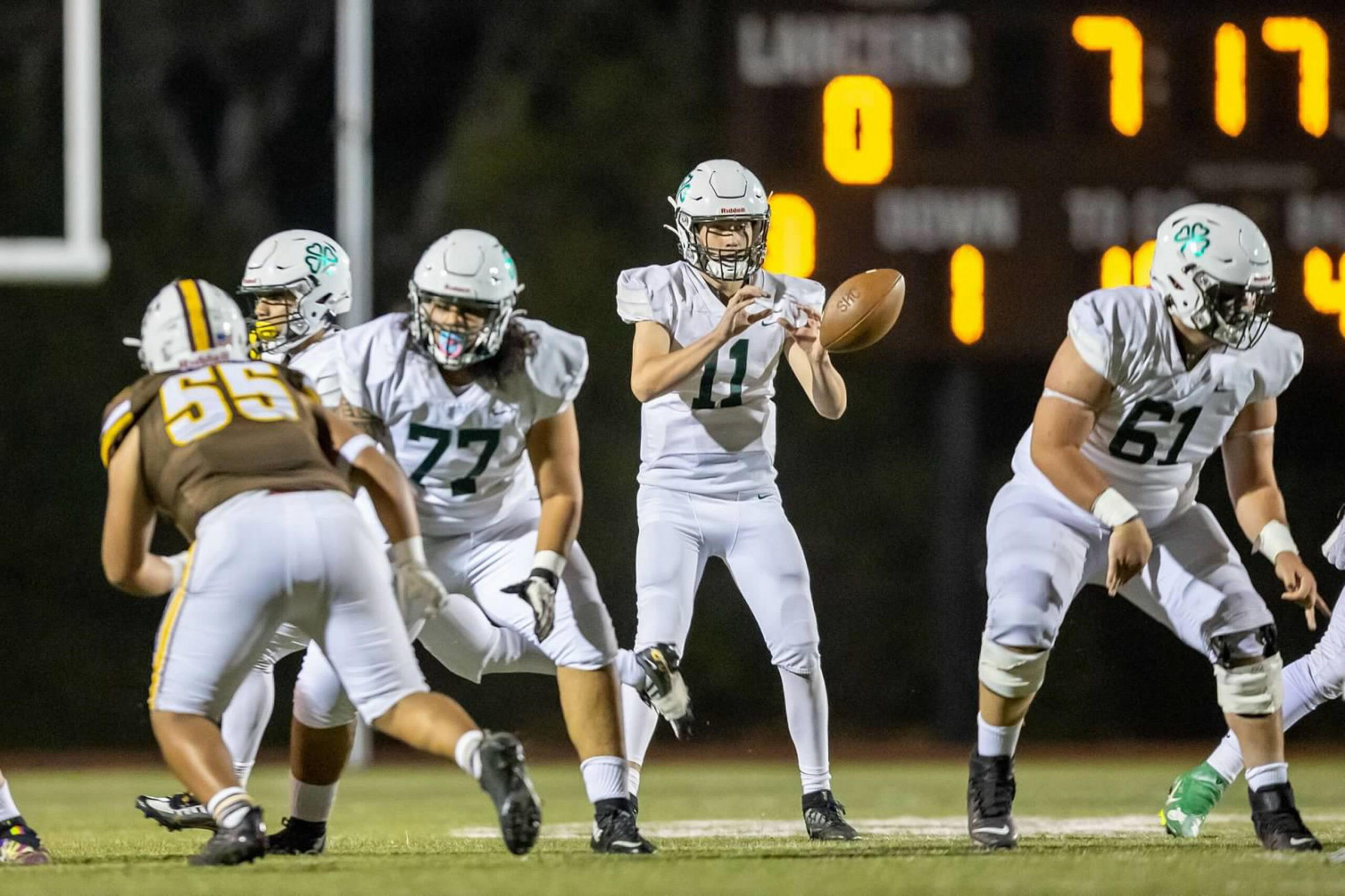 A football quarterback in white prepares to throw the ball while teammates block an approaching defender in brown and yellow.