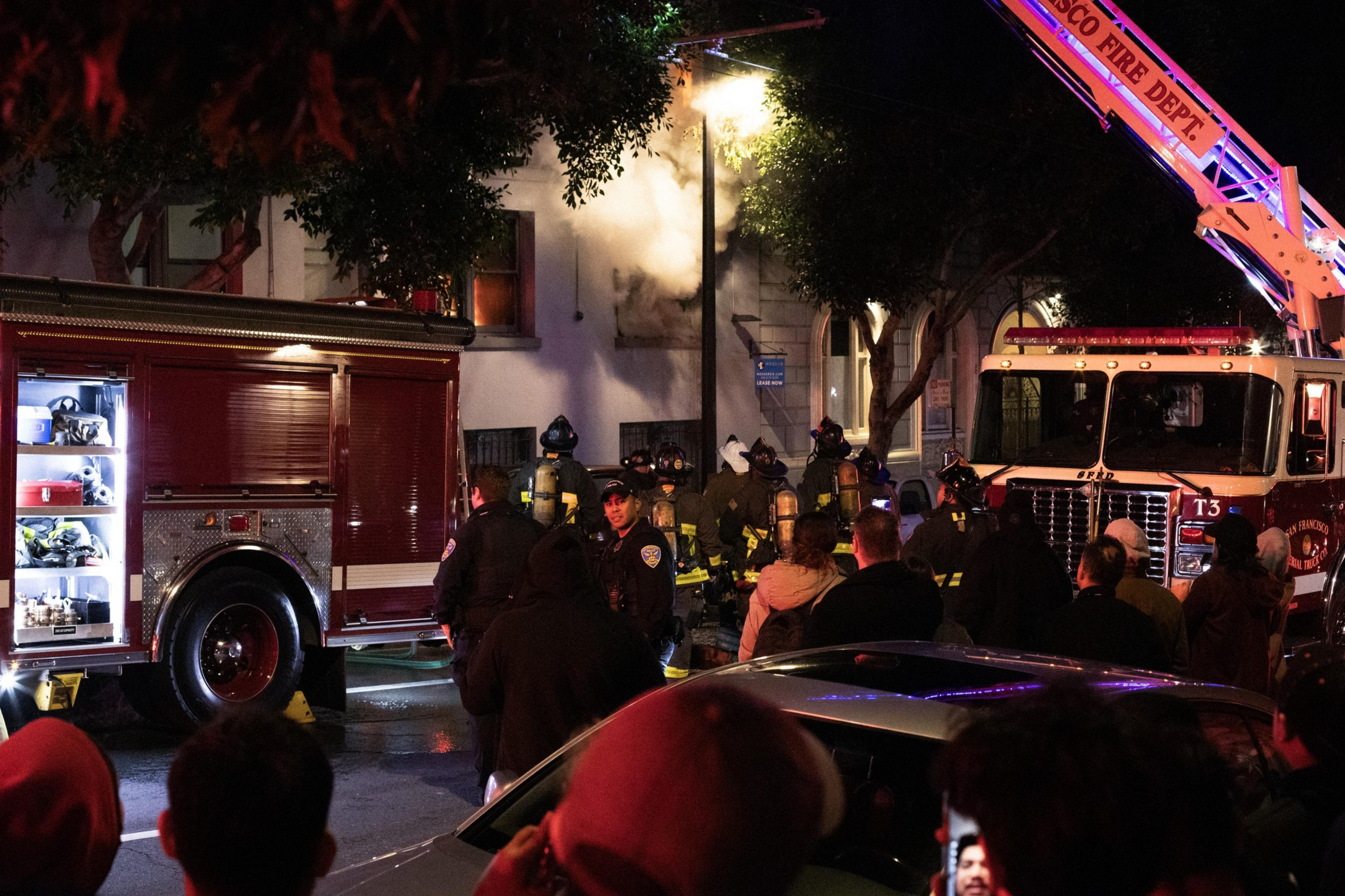 Firefighters watch smoke pour from a burned first-floor apartment building window.