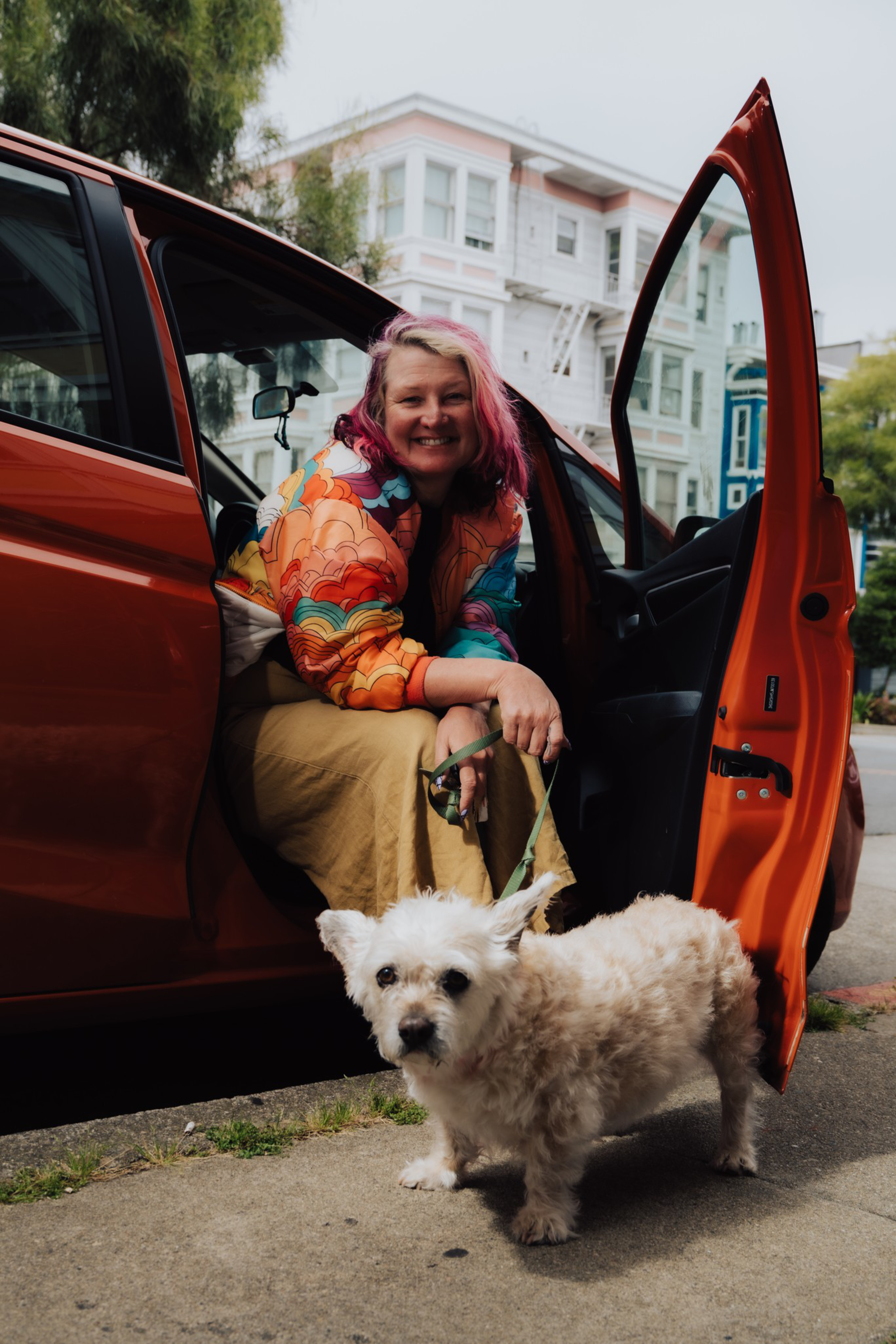 A smiling woman with pink hair sits in an open orange car door, holding the leash of a small, fluffy white dog standing on the sidewalk.