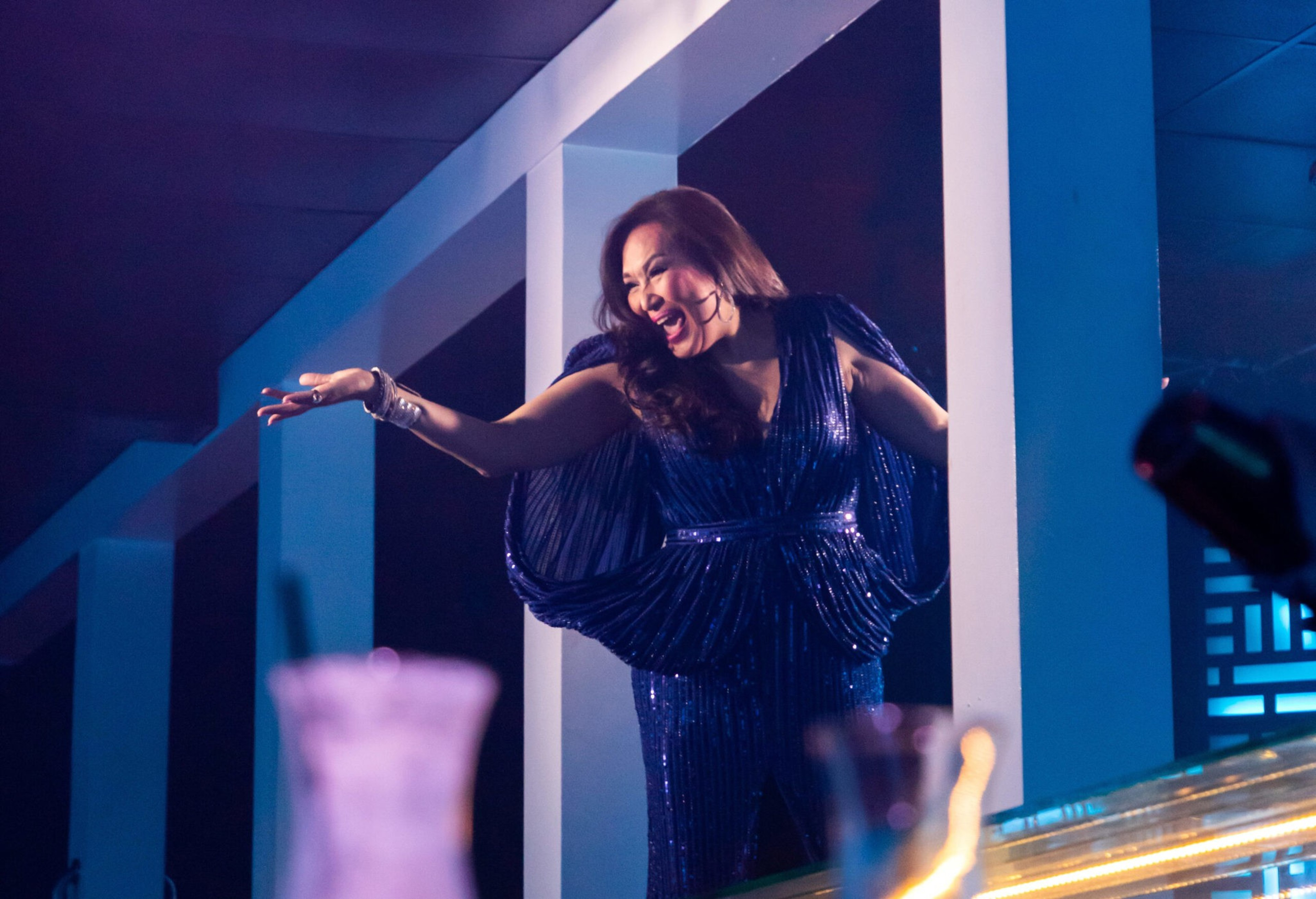 A woman in a shimmering dark blue gown extends her arm joyfully, illuminated by soft blue lighting in a modern indoor setting.