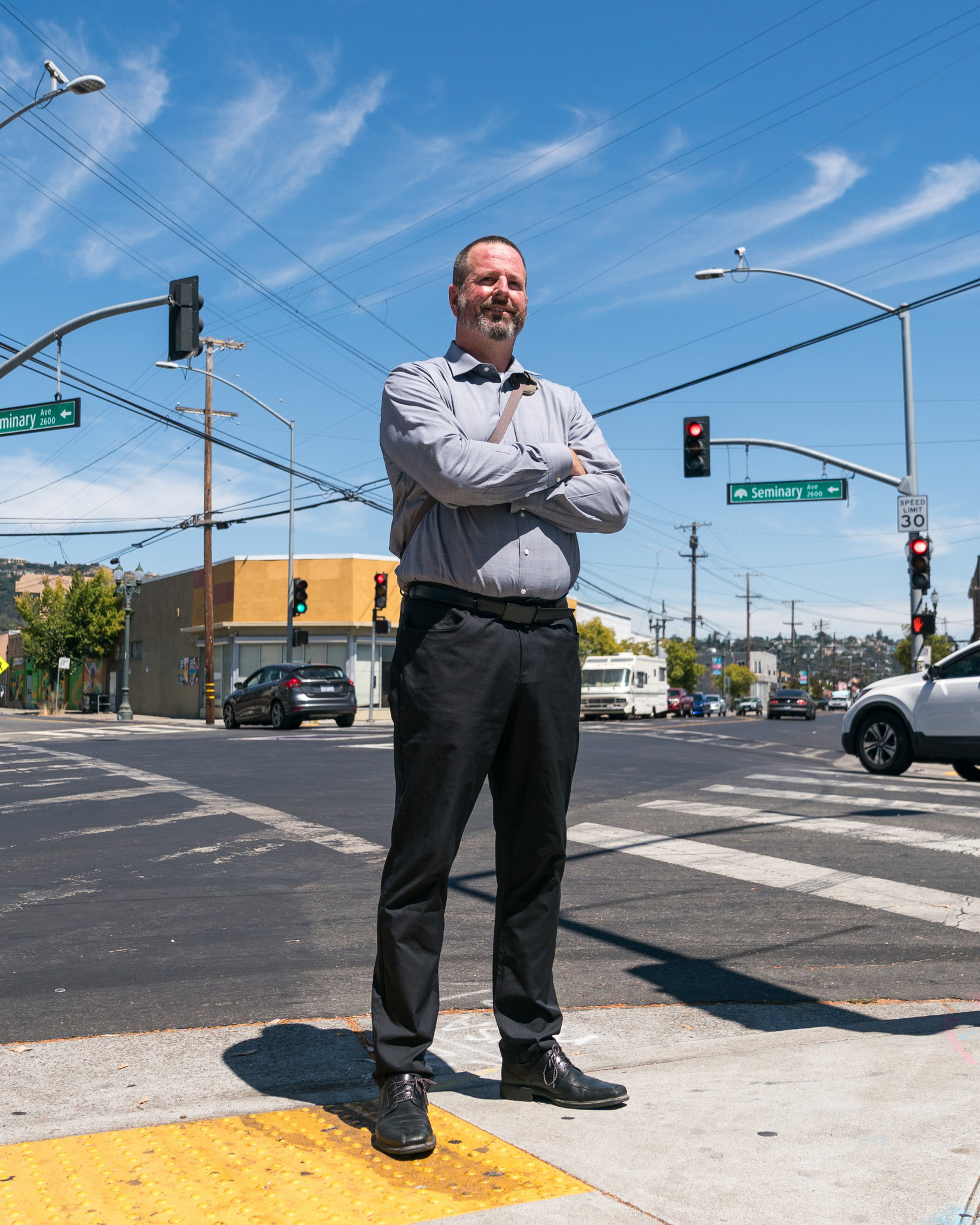 A man in a gray shirt and black pants stands confidently with arms crossed at a city intersection under a clear sky, with traffic signals and street signs visible.