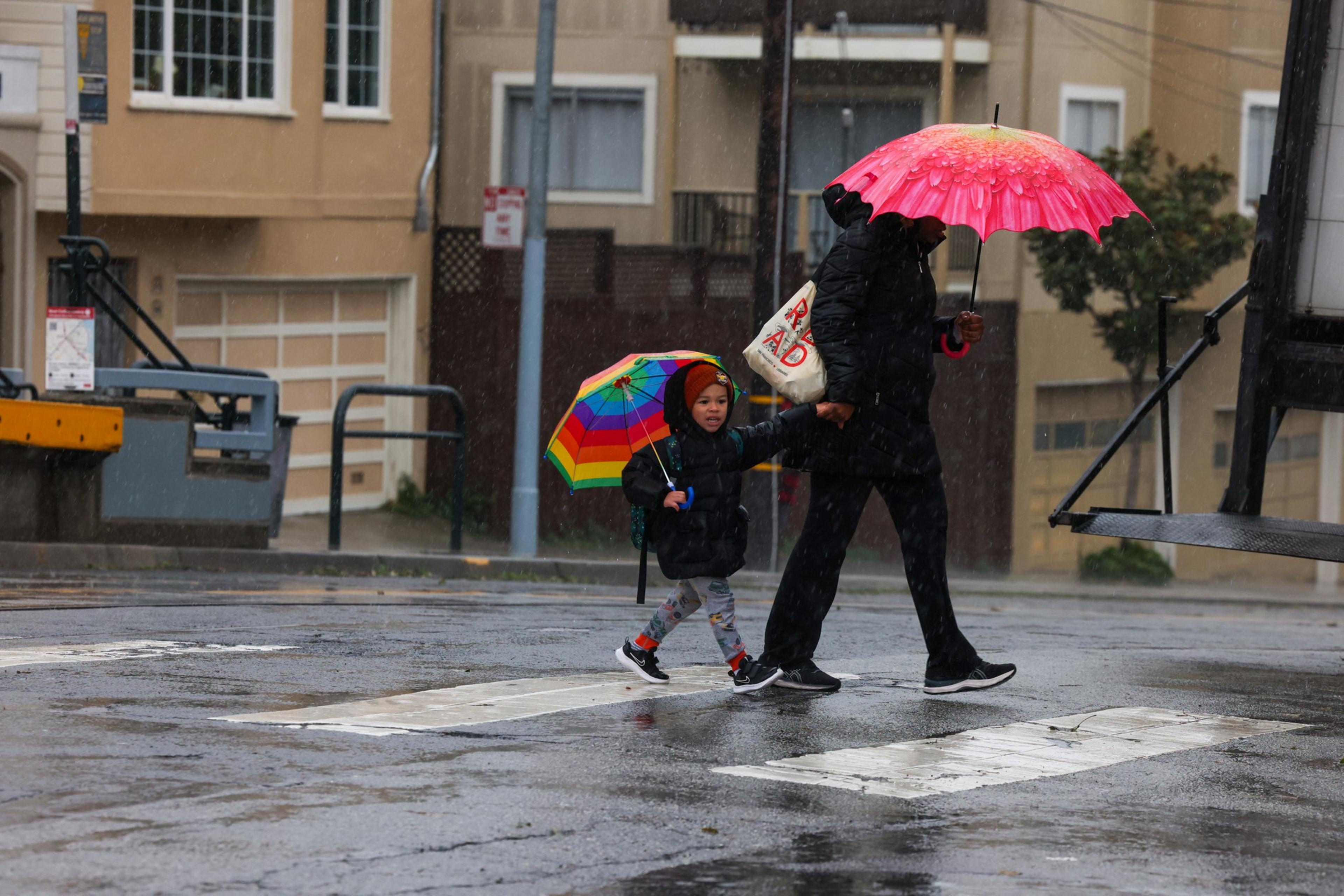 An adult and child walk across a wet street in the rain, holding hands; the adult has a pink umbrella, and the child carries a small rainbow umbrella.