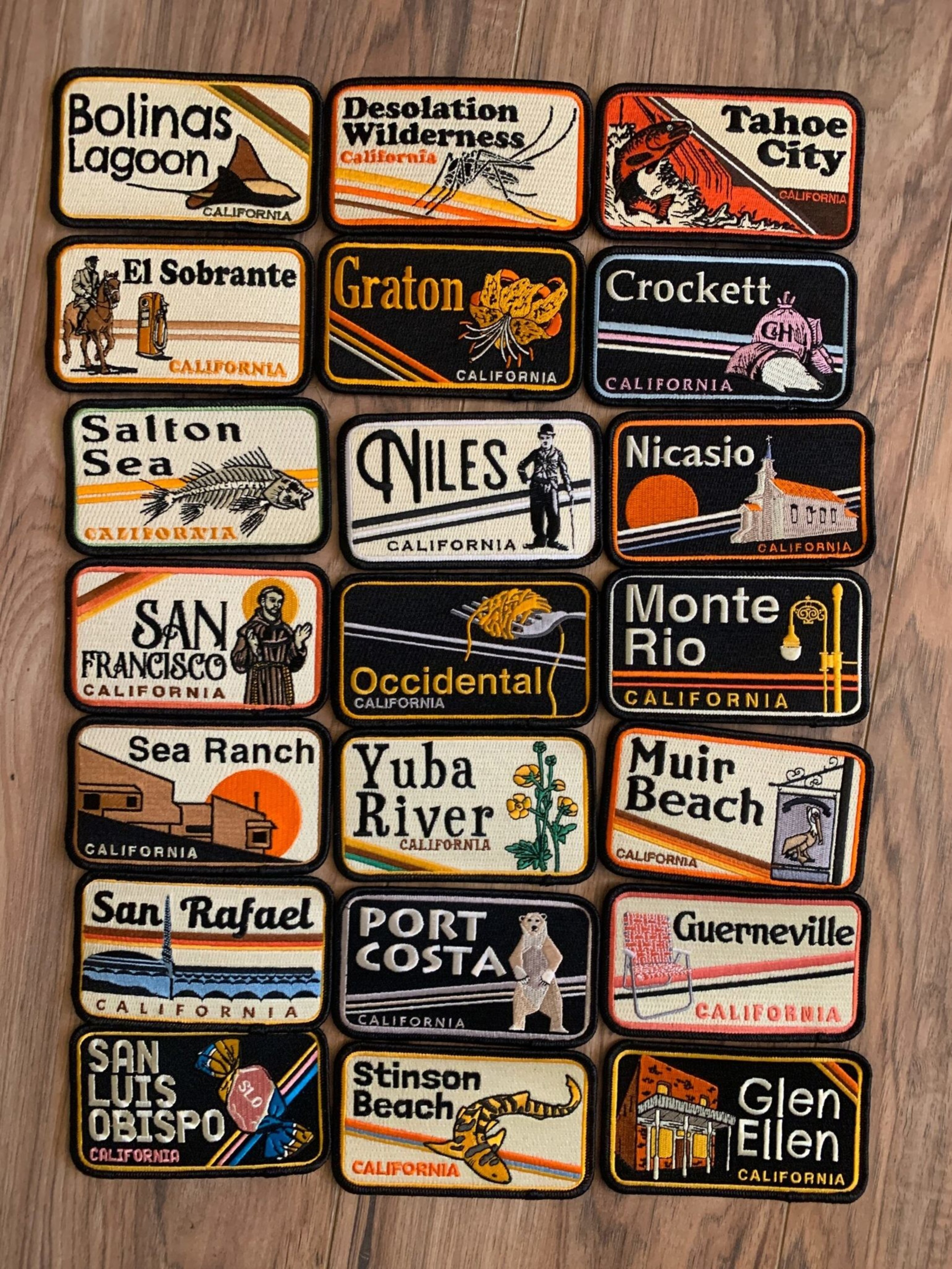 Collection of 20 embroidered patches featuring California locations, each with distinct illustrations and colorful text, arranged on a wooden surface.