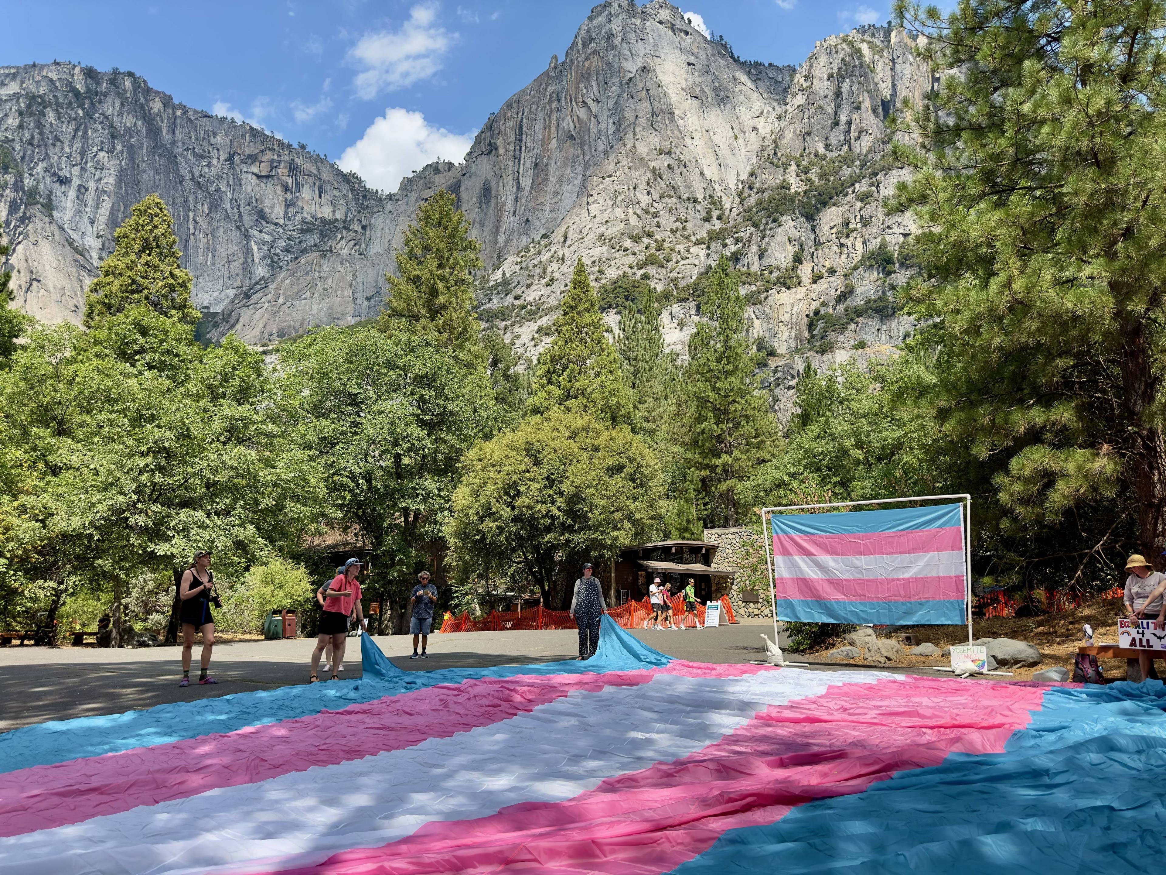 A large transgender pride flag is spread on the ground with people around it, set against a backdrop of tall trees and towering rocky cliffs.