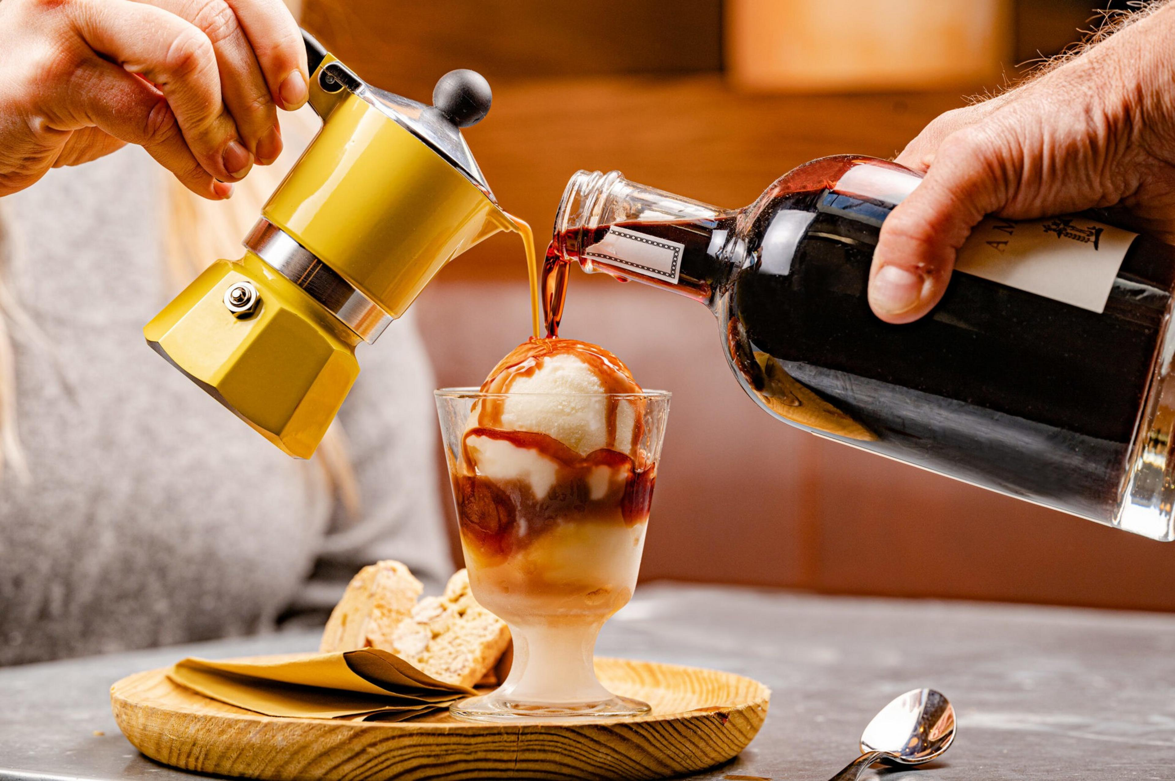 A glass with vanilla ice cream is topped with espresso poured from a small yellow pot and dark liqueur from a bottle, placed on a wooden plate with biscotti.