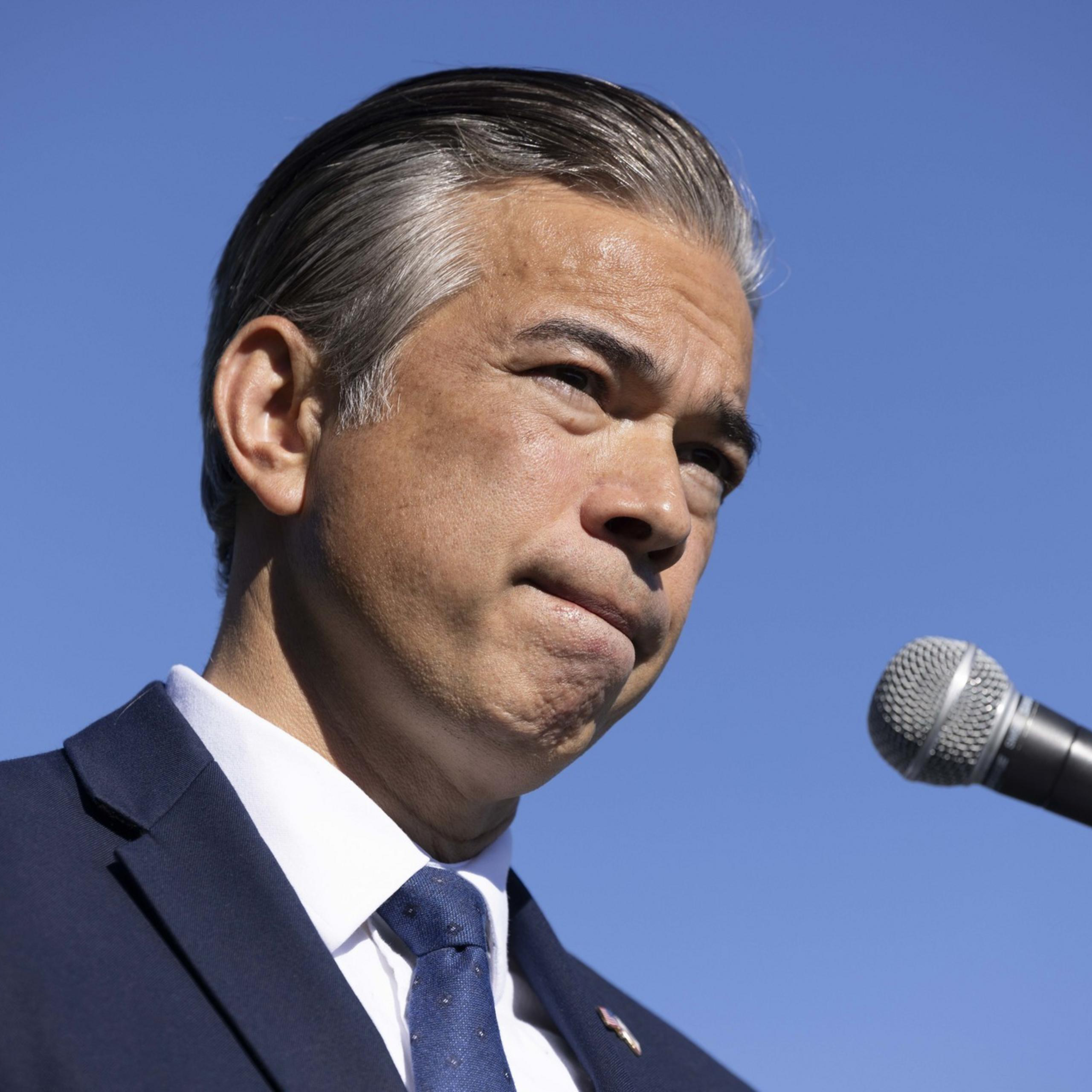 A man in a suit stands against a clear blue sky, speaking into a microphone with a focused expression.