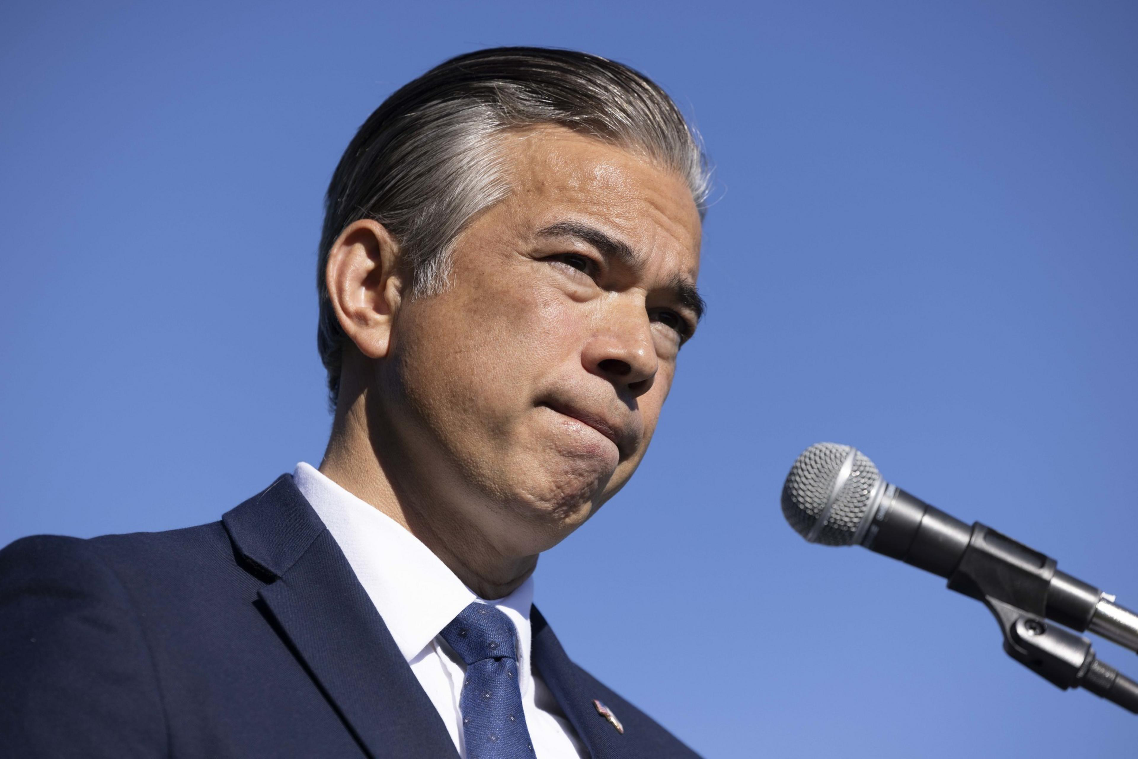 A man in a suit stands against a clear blue sky, speaking into a microphone with a focused expression.