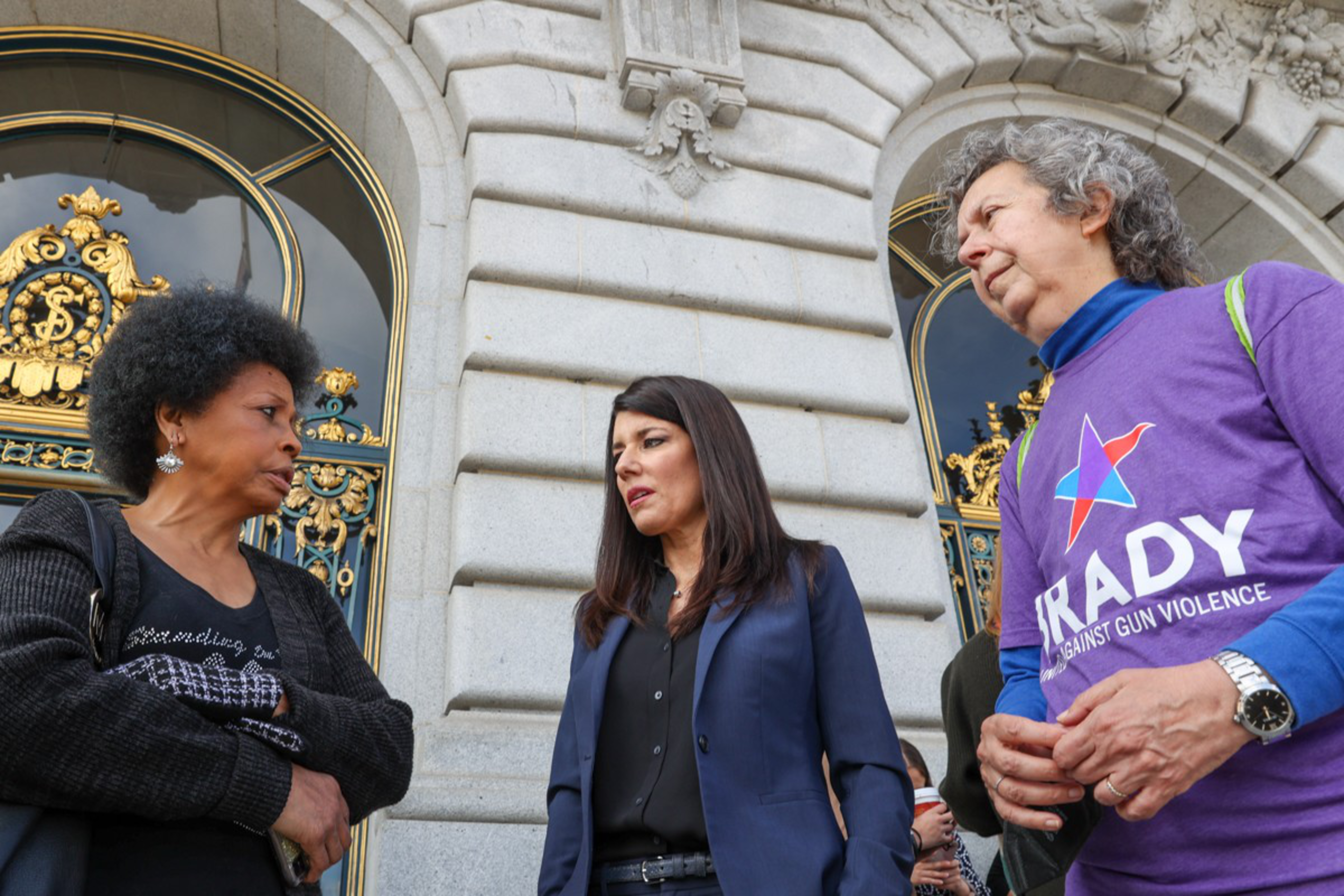 Three people stand outside an ornate, arched building entrance, having a conversation. One wears a Brady campaign shirt, another a suit, and the third a sweater.