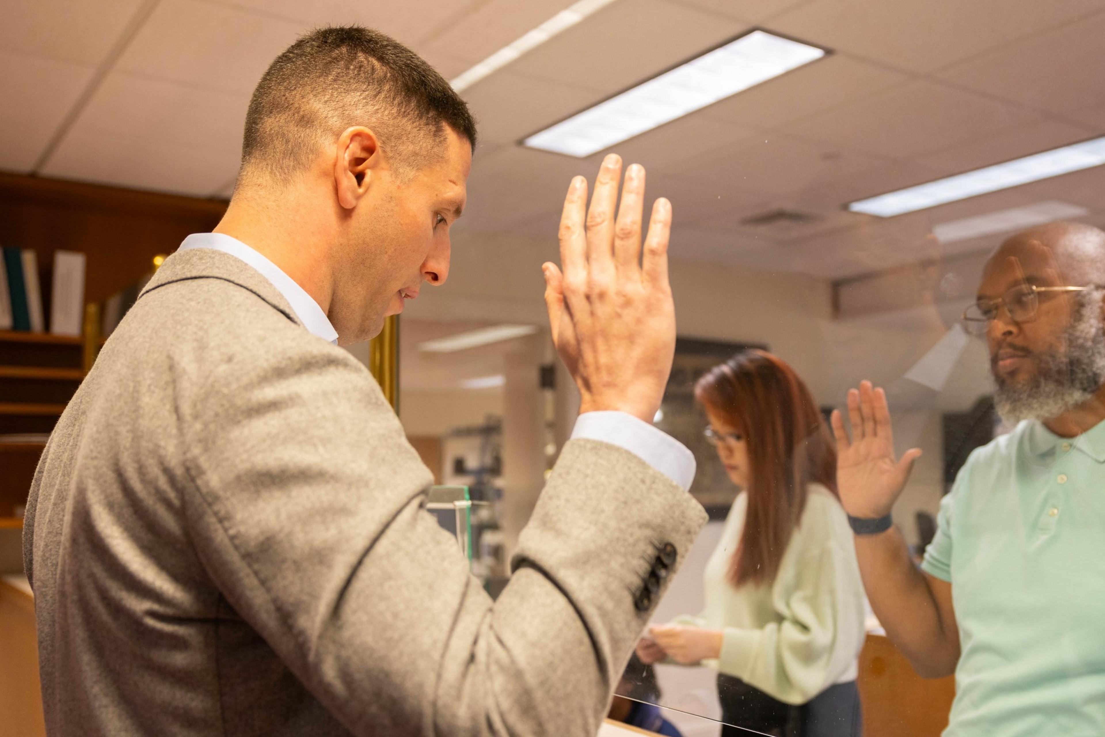 Jay Donde raises his right hand in the direction of another man raising his right with a pane of glass between them as San Francisco City Hall Board of Elections Department.