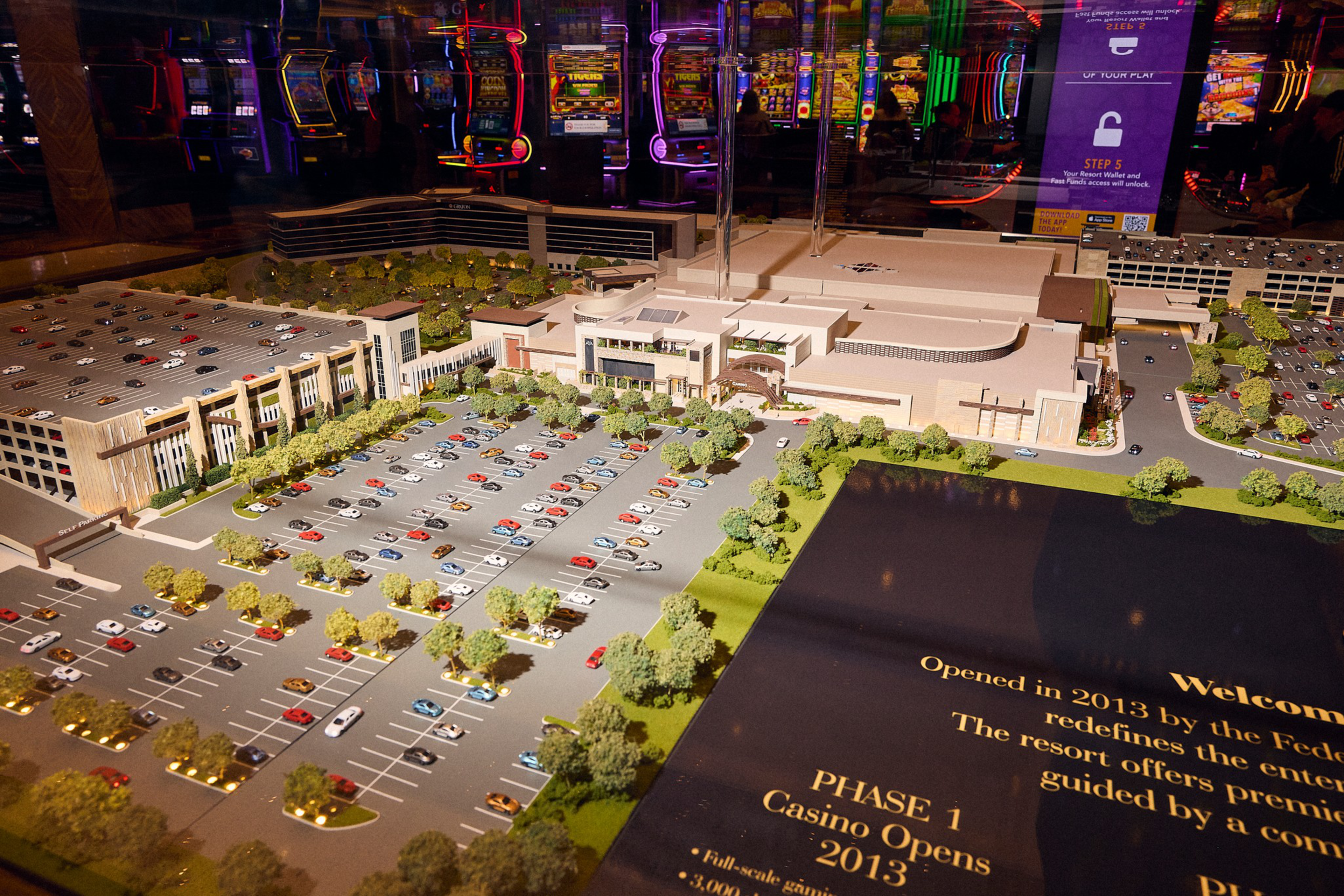 A detailed scale model of a casino complex includes multiple parking lots filled with miniature cars, surrounding trees, and a large main building.