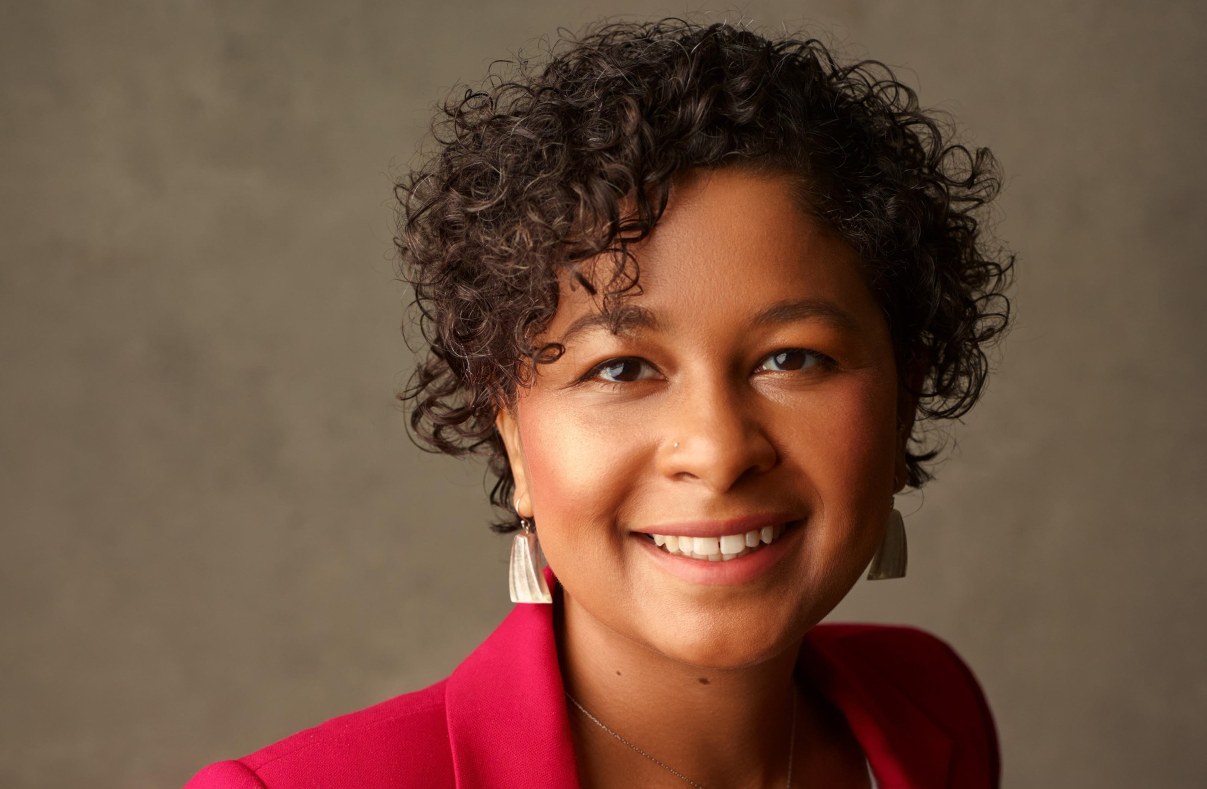 A person with short curly hair smiles warmly, wearing a red blazer and light-colored earrings against a plain neutral background.