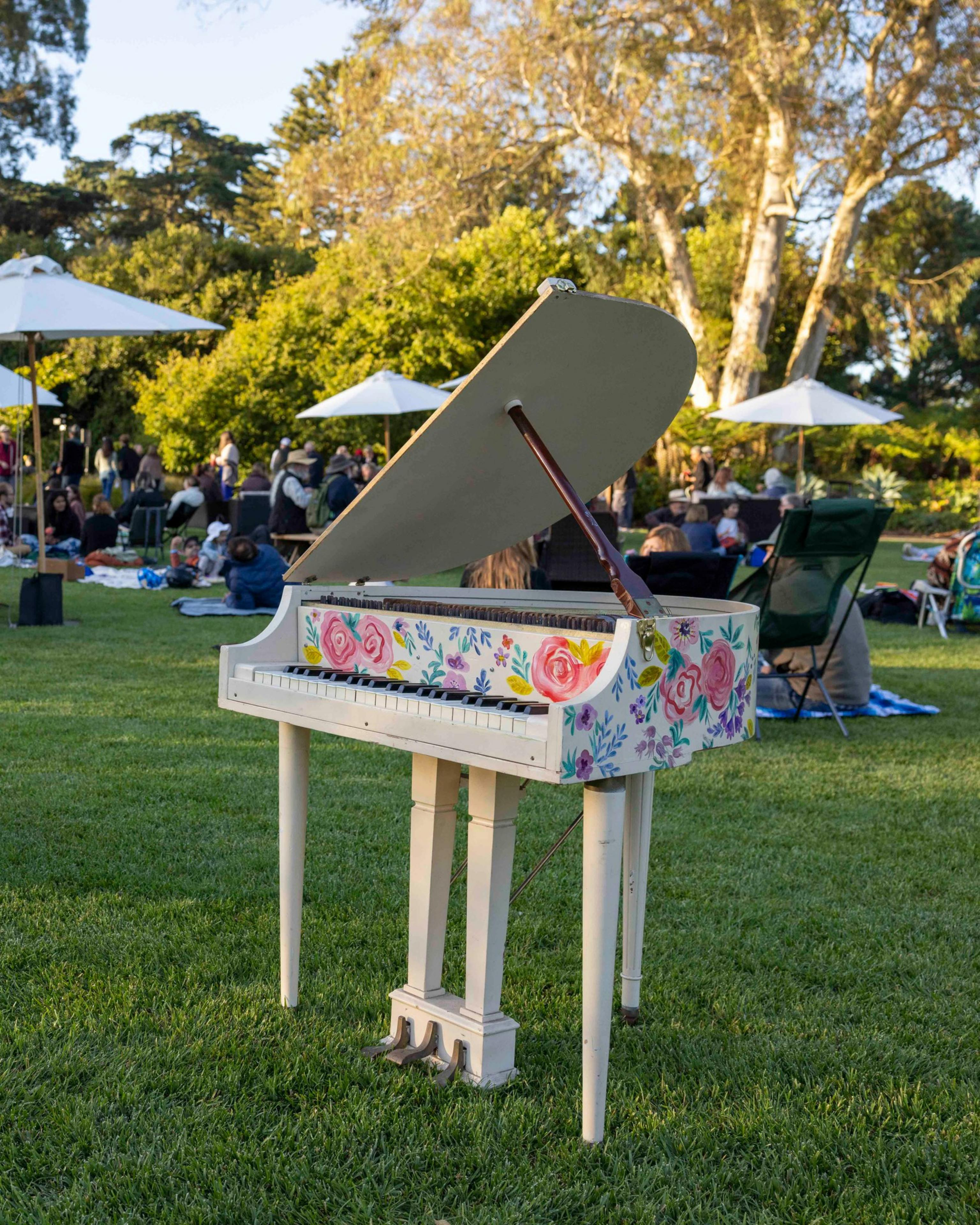 In a park, a small white grand piano with colorful flower designs stands on the grass. People are gathered and seated, with white umbrellas and trees in the background.