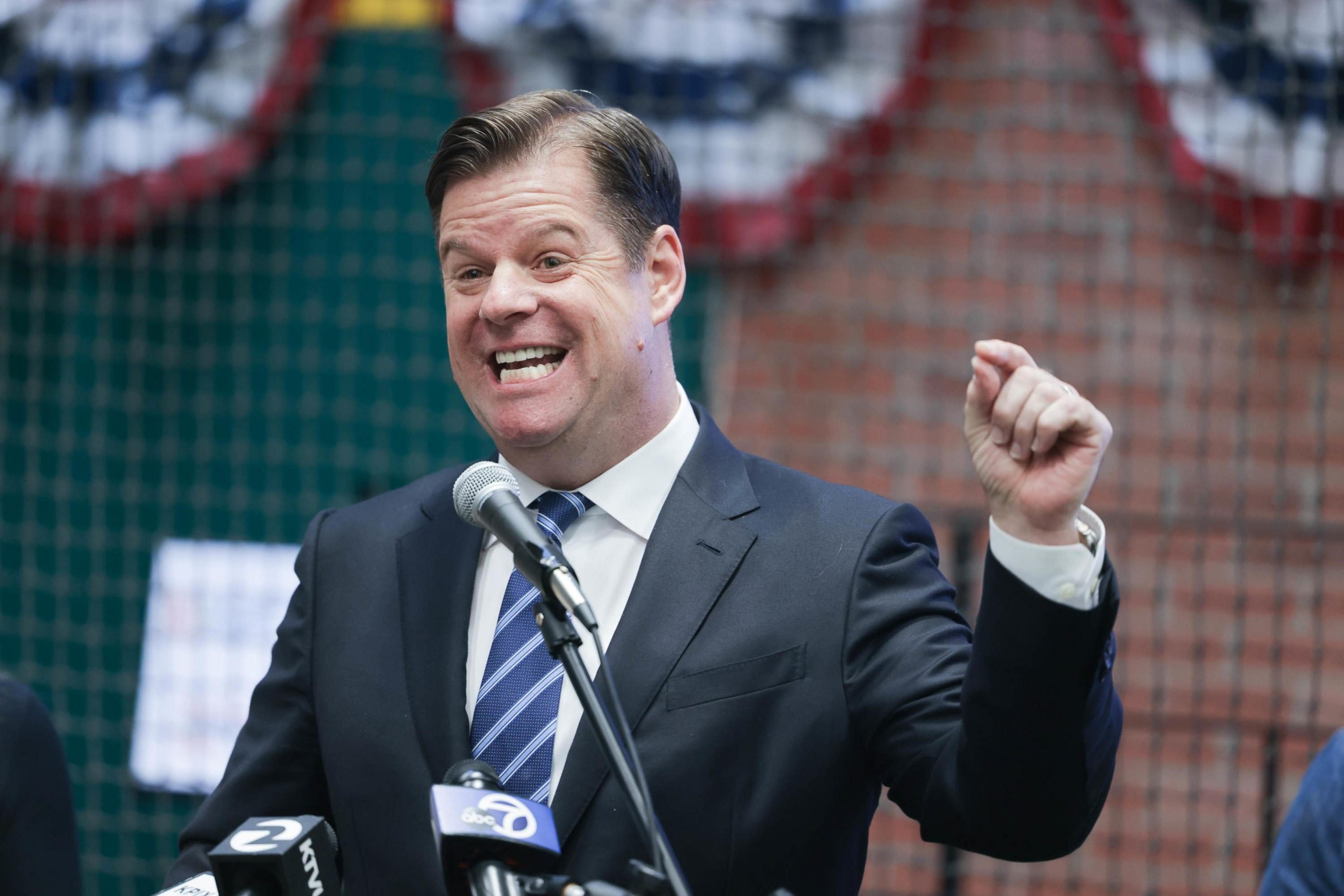 A man in a suit is speaking at a podium with microphones, gesturing with a raised fist, showing enthusiasm or making a point.