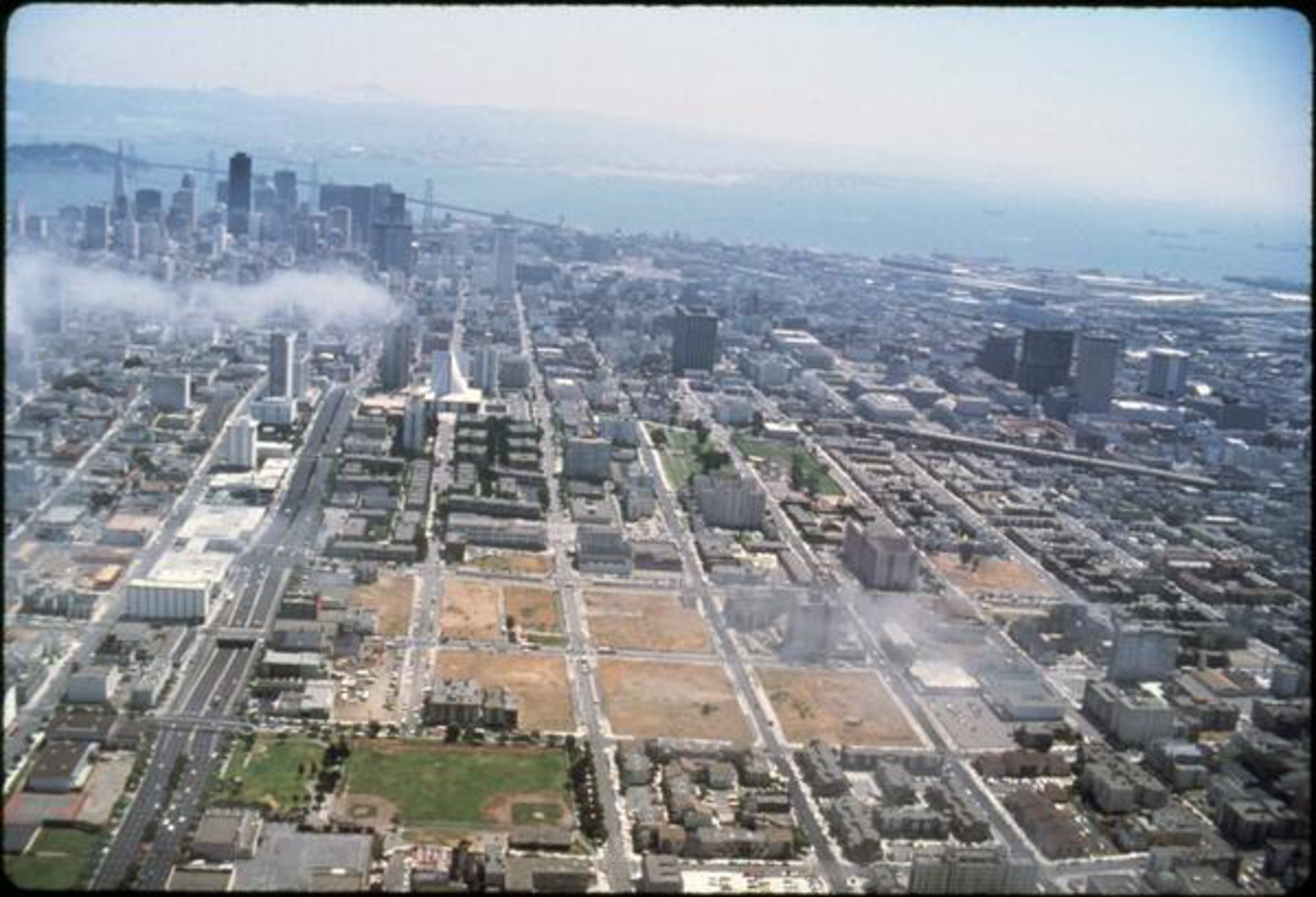 Aerial view of a cityscape with dense buildings, streets in a grid pattern, and a hazy skyline.