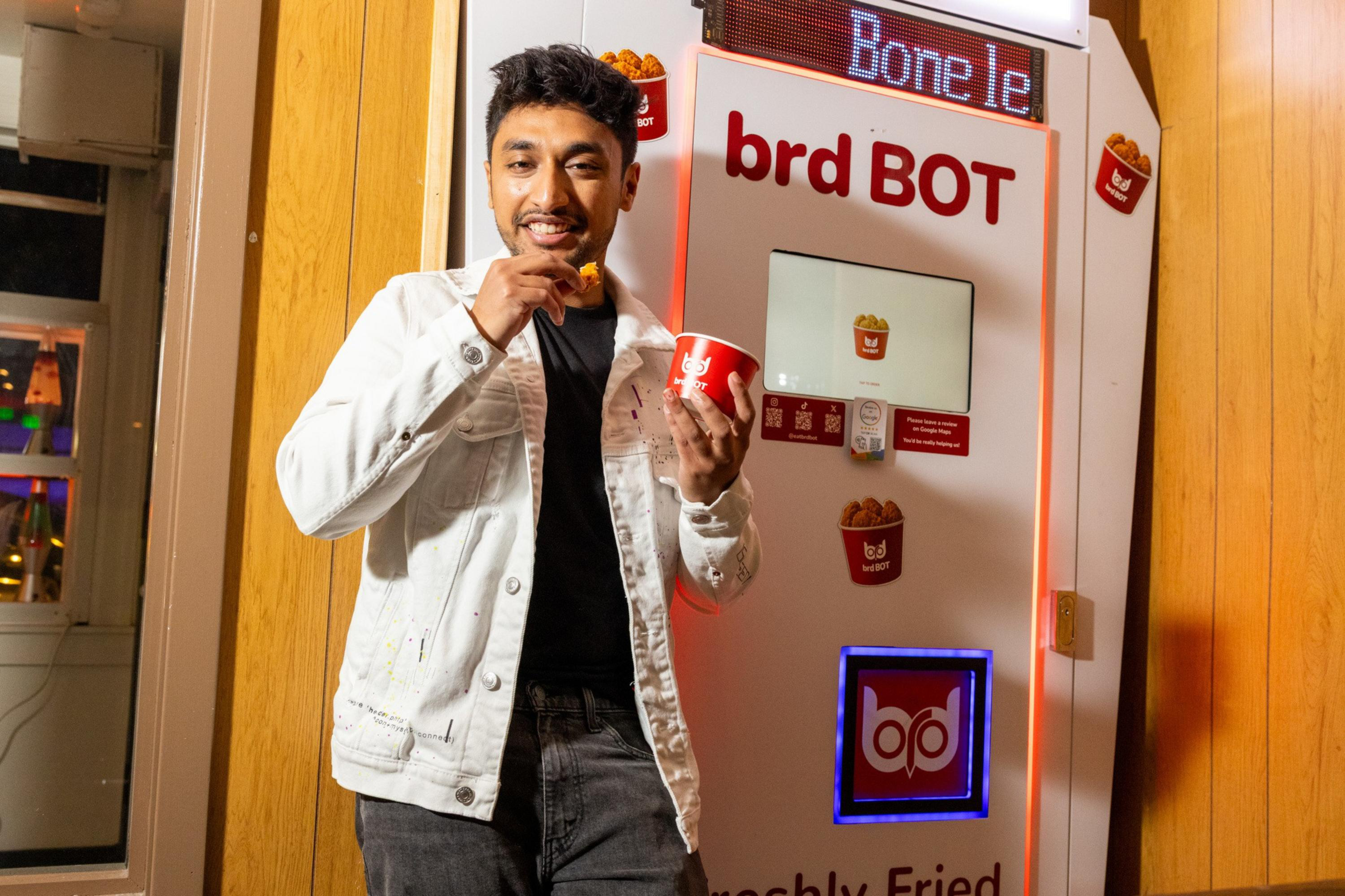 A man in a white jacket eats fried chicken from a red cup while standing next to a food vending machine labeled “brd BOT.”