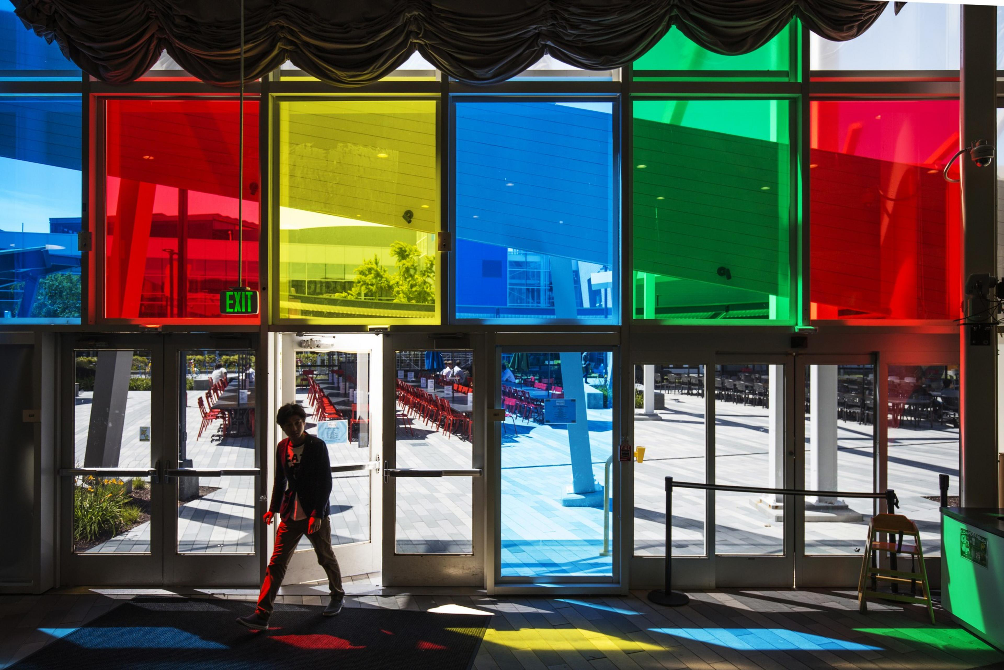 A person walks inside through glass doors with colorful window panels casting red, yellow, blue, and green hues on the ground. An "Exit" sign hangs above.