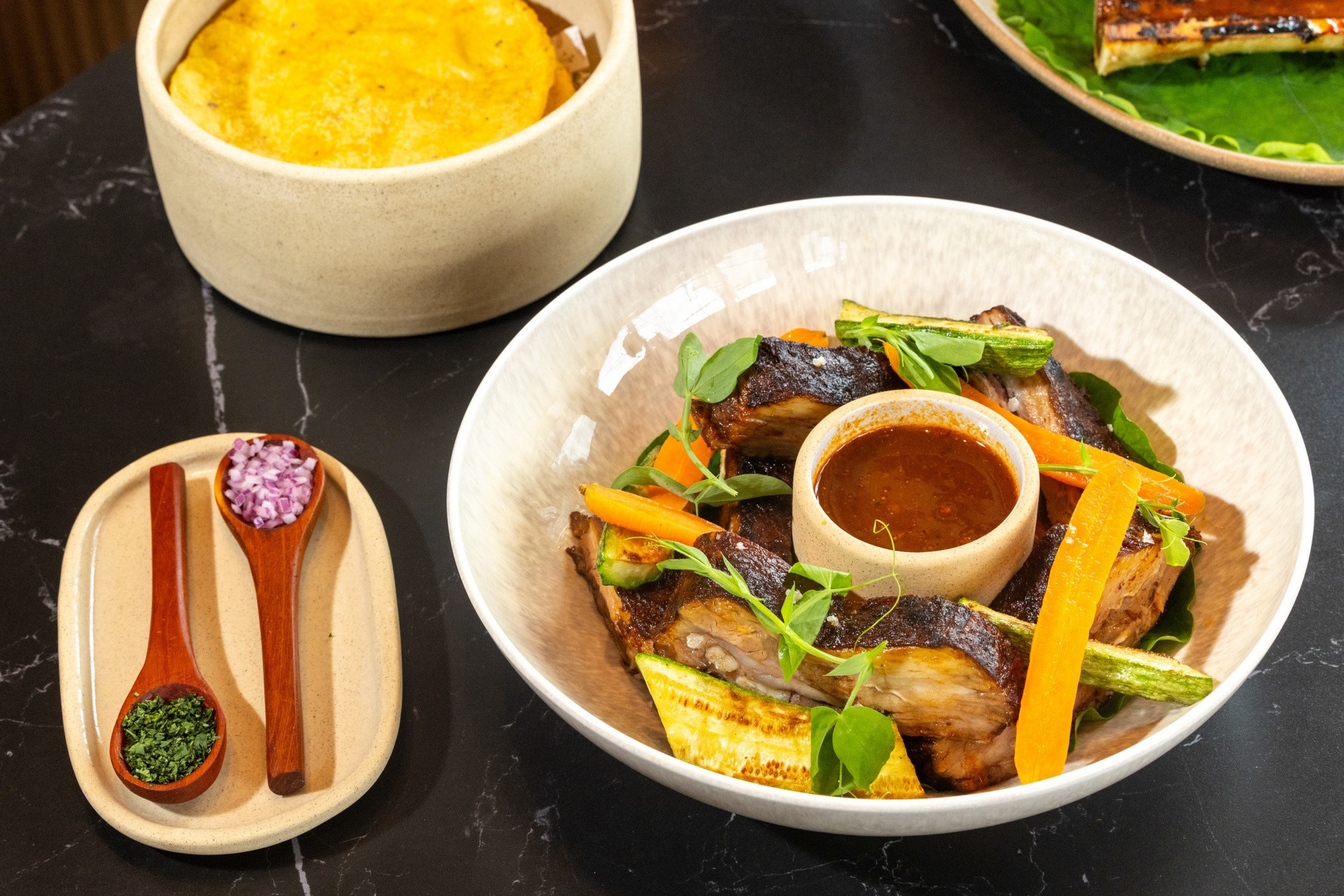 Grilled pork ribs served with carrots, asparagus, greens, a bowl of dipping sauce, finely chopped onions and parsley, and a bowl of tortillas.