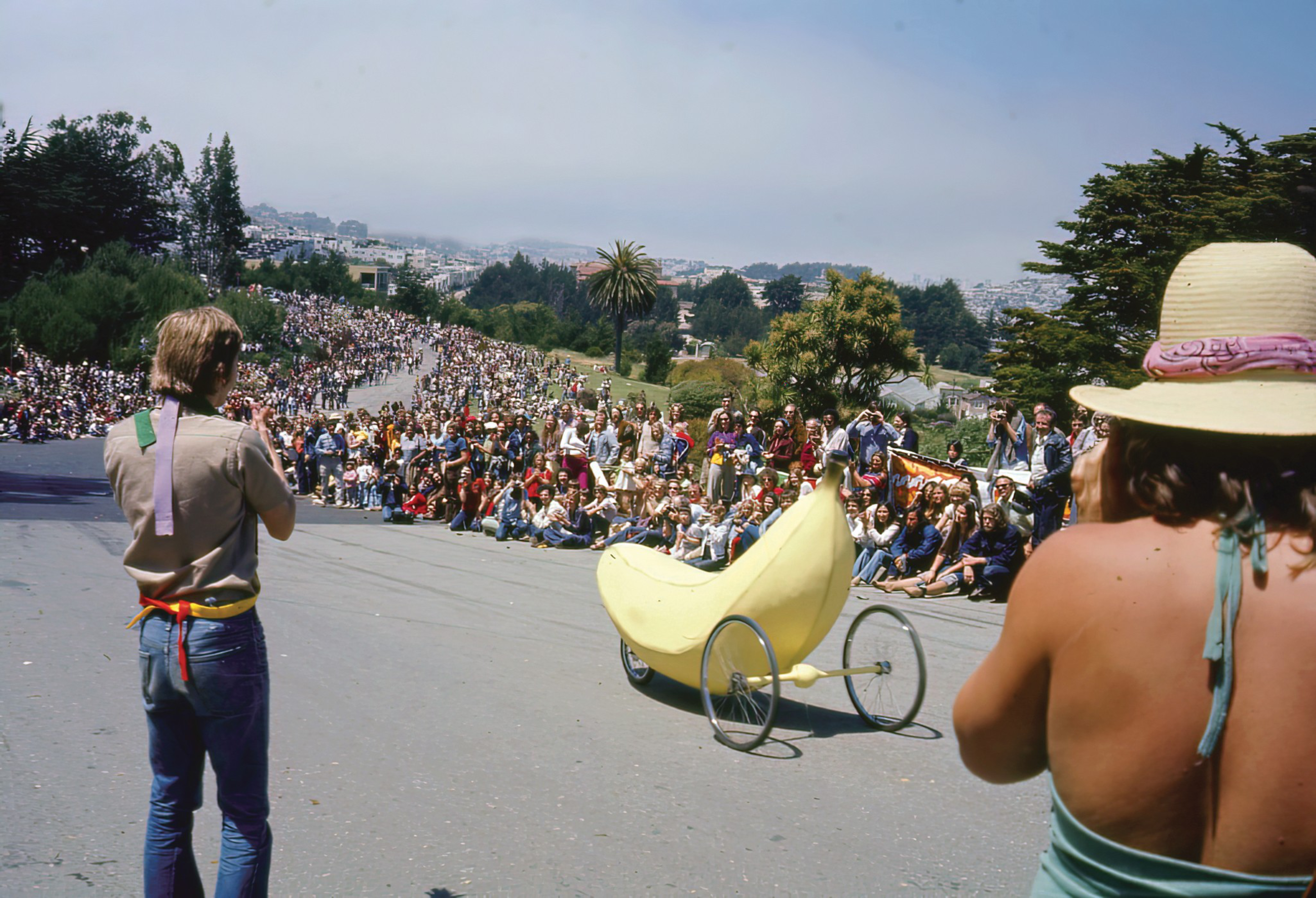 A large crowd lines a winding road as a person pedals a banana-shaped vehicle while two spectators take photos.