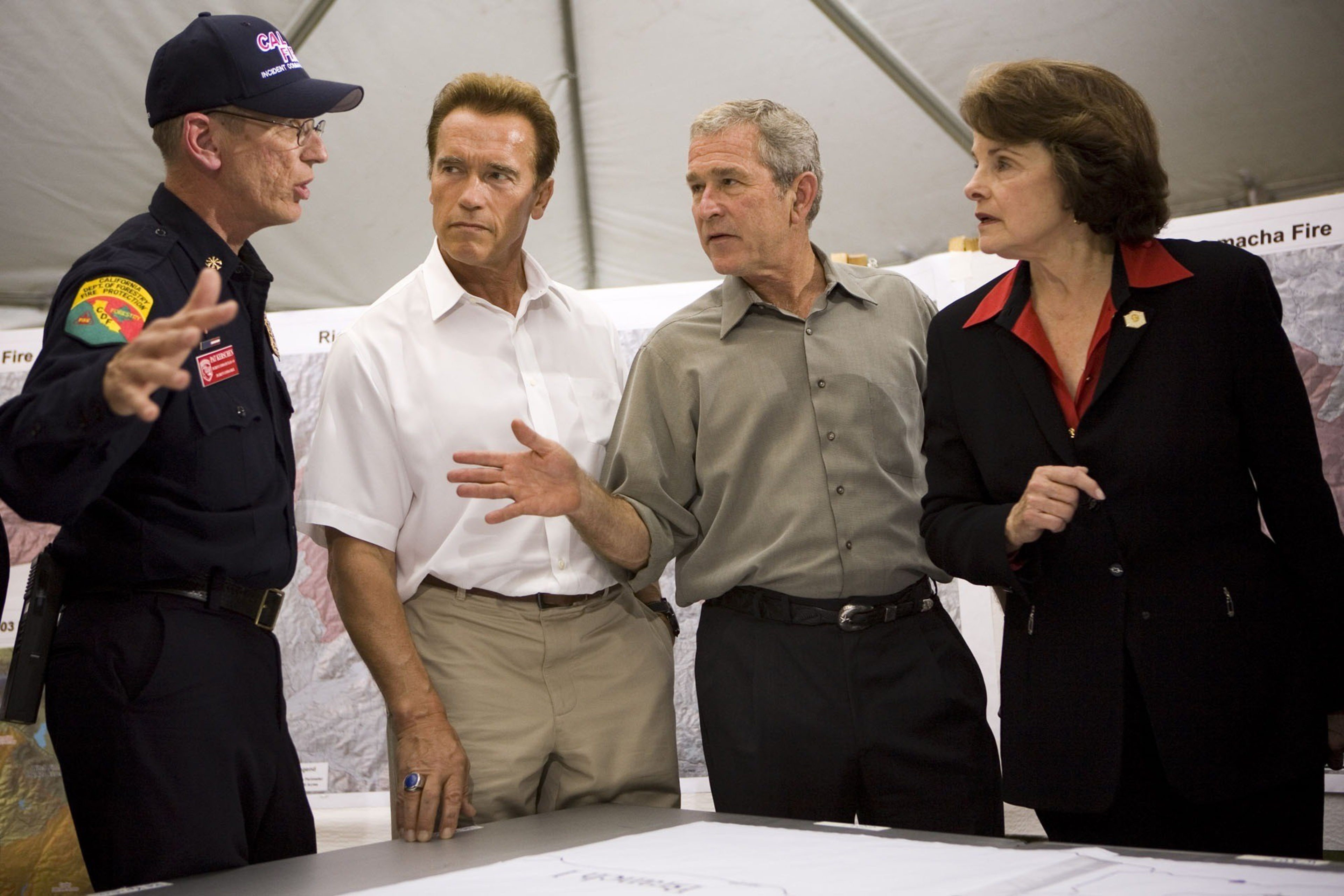 Four adults stand around a table, engaged in discussion; one man in uniform gestures while the others listen attentively, surrounded by maps and a tent.