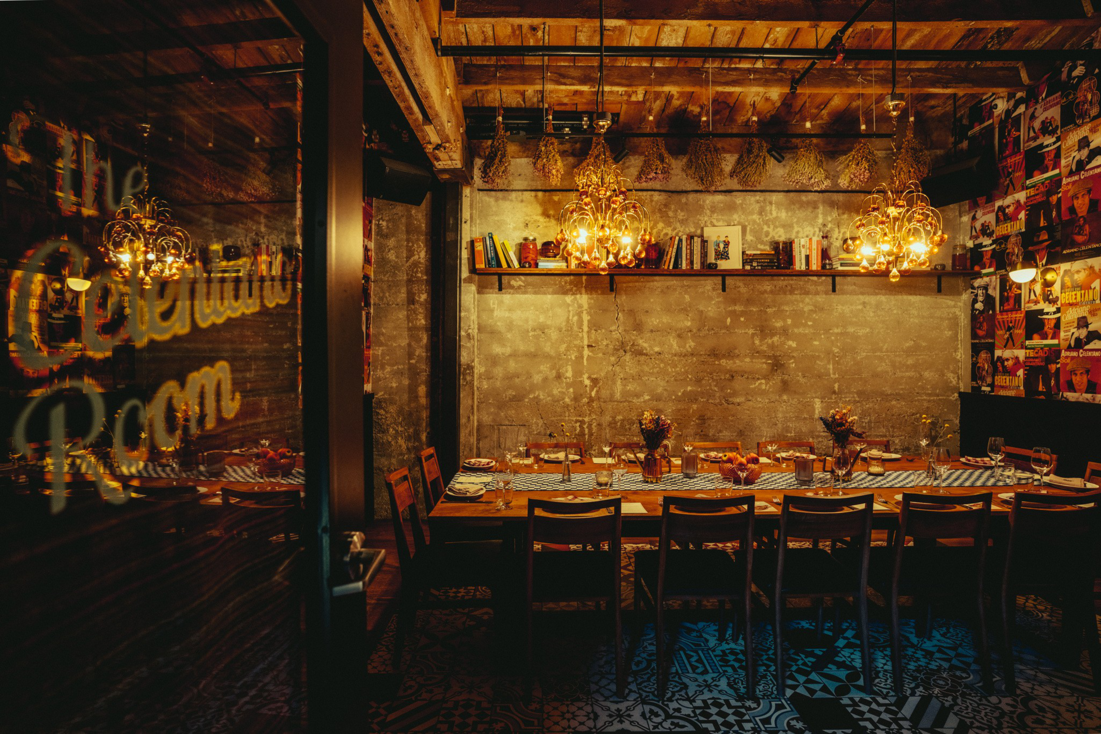 A warmly lit dining room features a long wooden table set for a meal, surrounded by chairs. Chandeliers and books add a cozy ambiance, with a rustic-industrial decor.